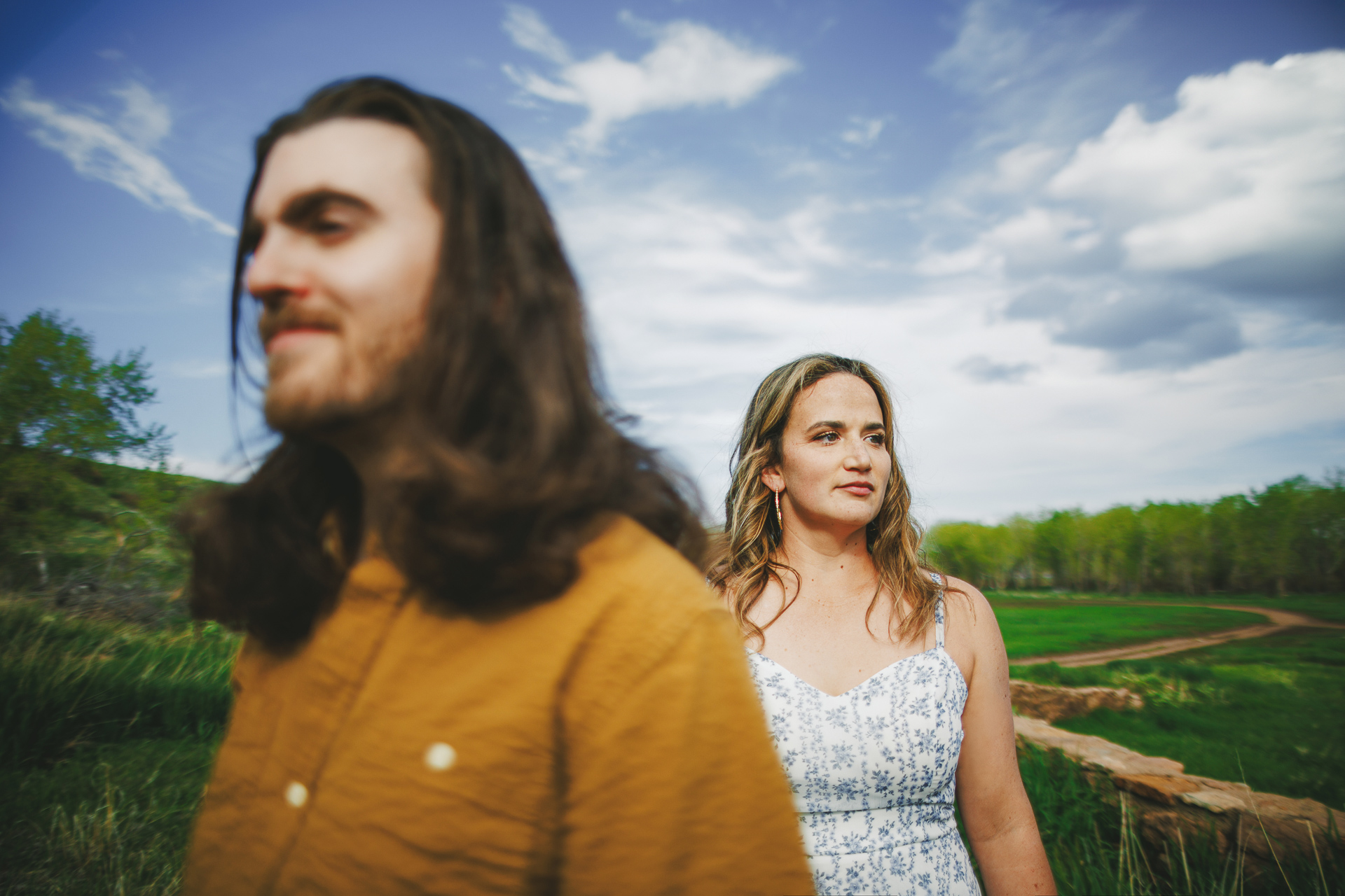 A man and a woman stand outdoors with a scenic background of green fields and trees under a partially cloudy sky. The man is in the foreground, slightly out of focus as if captured by auto draft, while the woman is in the background, in focus.