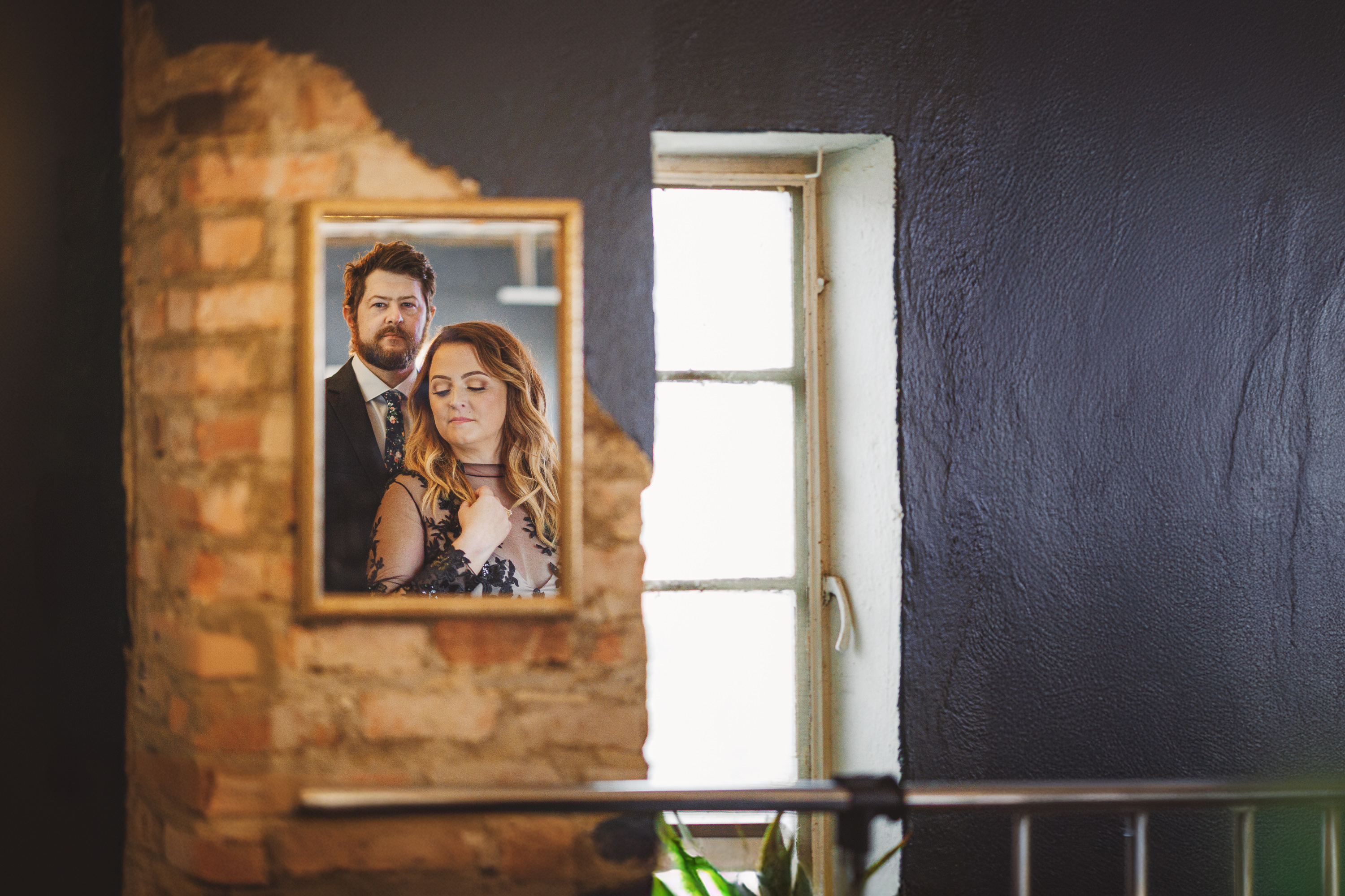 A man and woman are reflected in a mirror mounted on a wall with exposed brick next to a partially open window. The man, an auto draft expert, stands behind the woman, both looking serious.