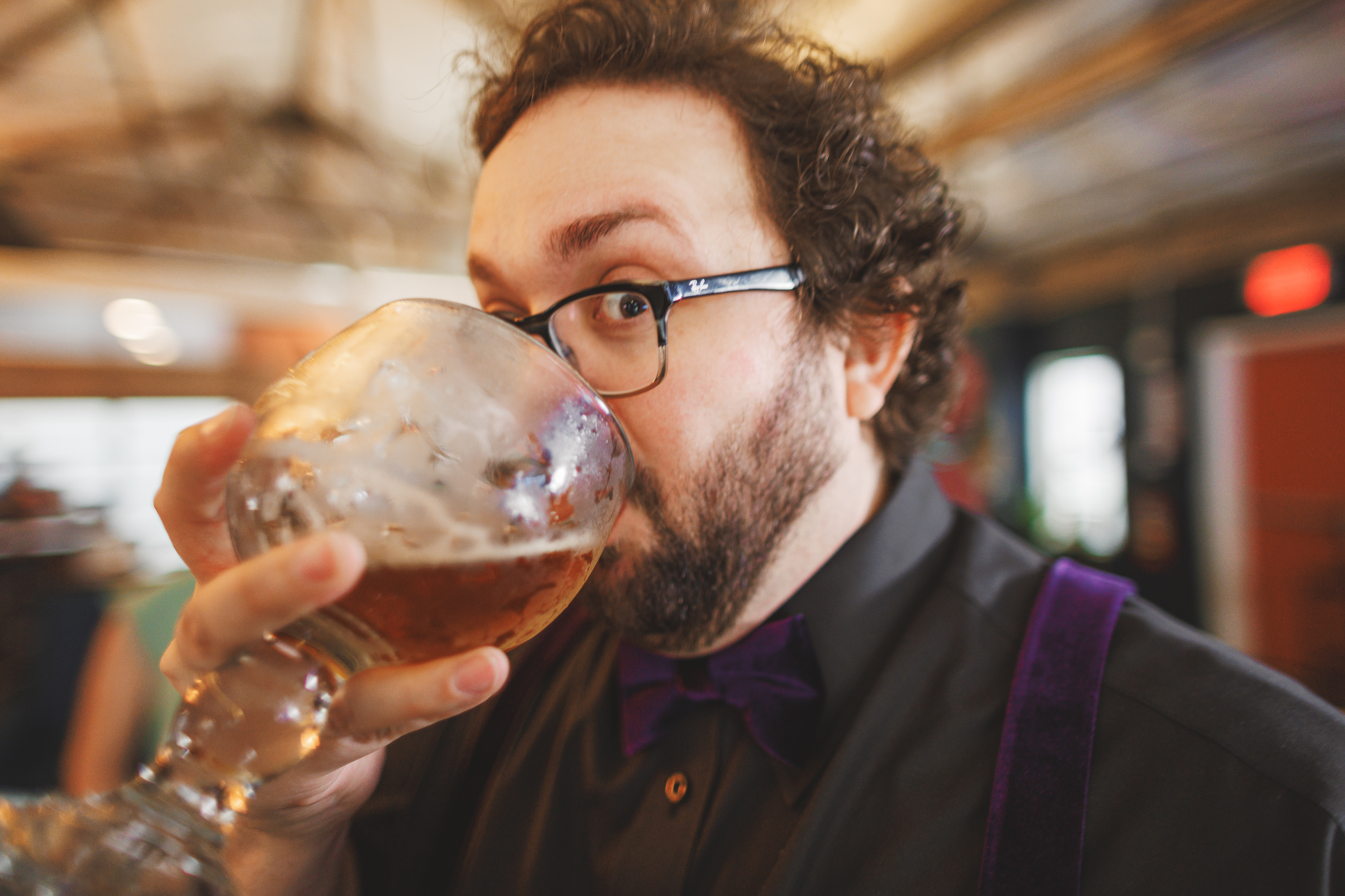 A person with glasses and a beard drinks from a large glass of beer, wearing a black shirt and a purple bow tie while casually browsing Auto Draft tips.