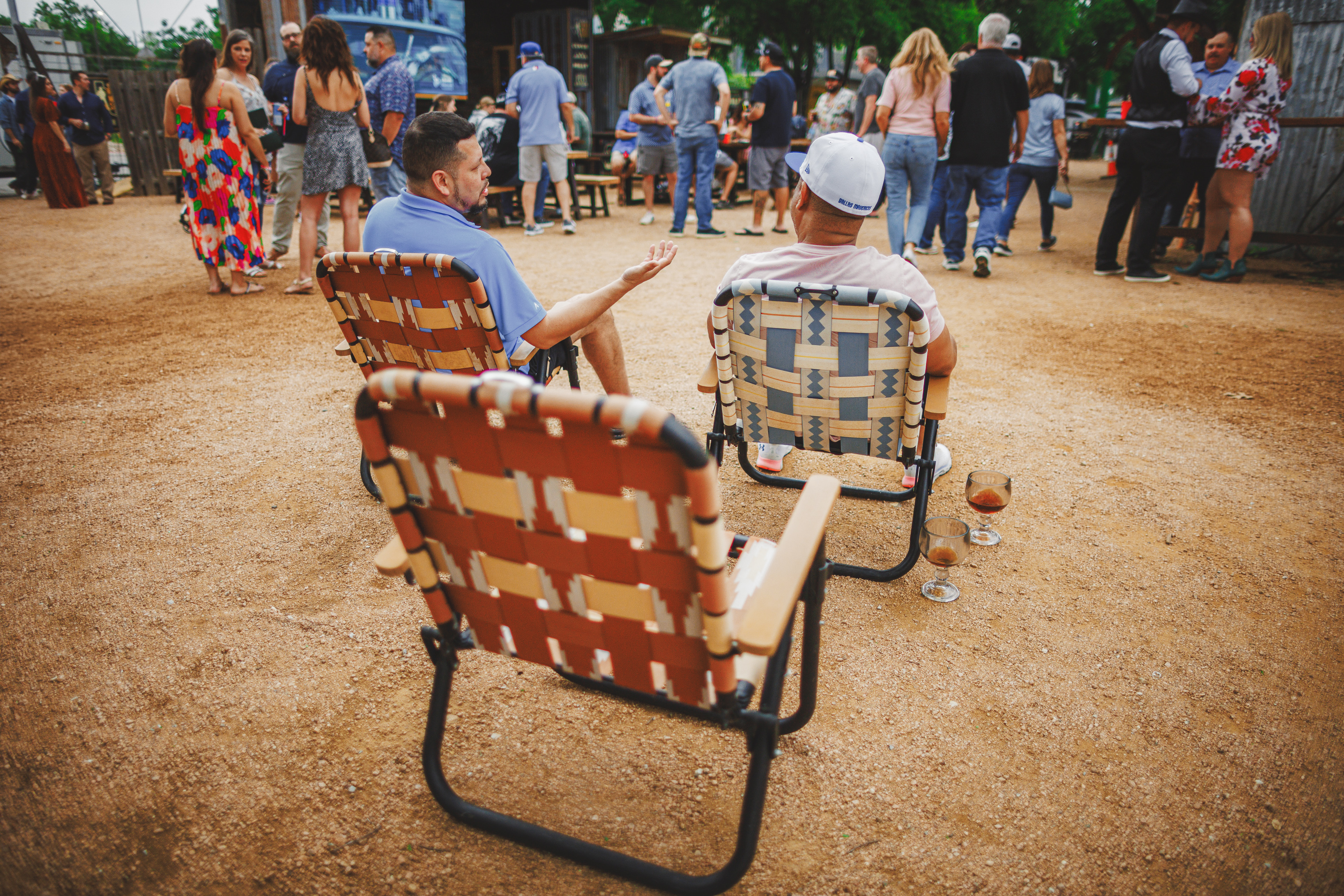 A group of people socialize outdoors. Two men sit on woven lawn chairs, their drinks on the ground beside them, engaging in conversation about the latest Auto Draft features. Other people are standing and mingling in the background.