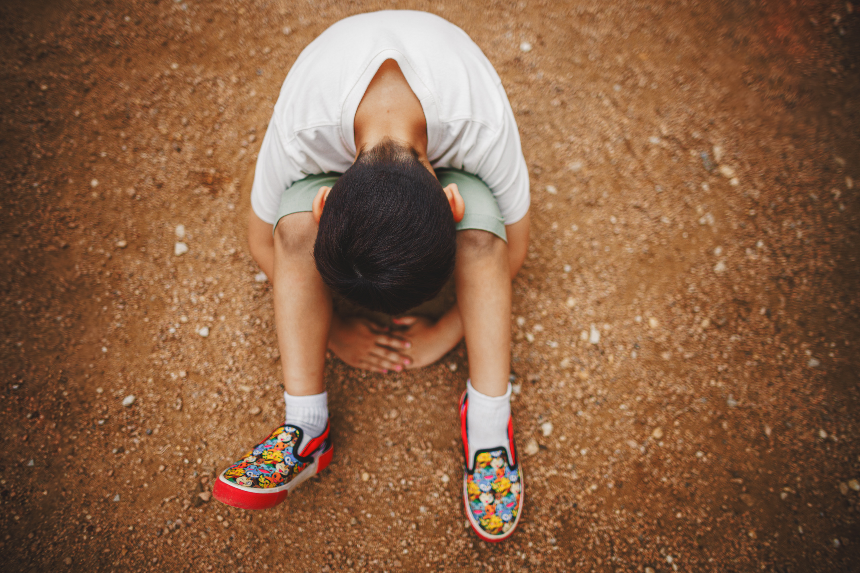 A child sits on the ground with their head down, arms wrapped around their knees, and wearing colorful shoes, like a whimsical auto draft of innocence amidst the world's hustle.