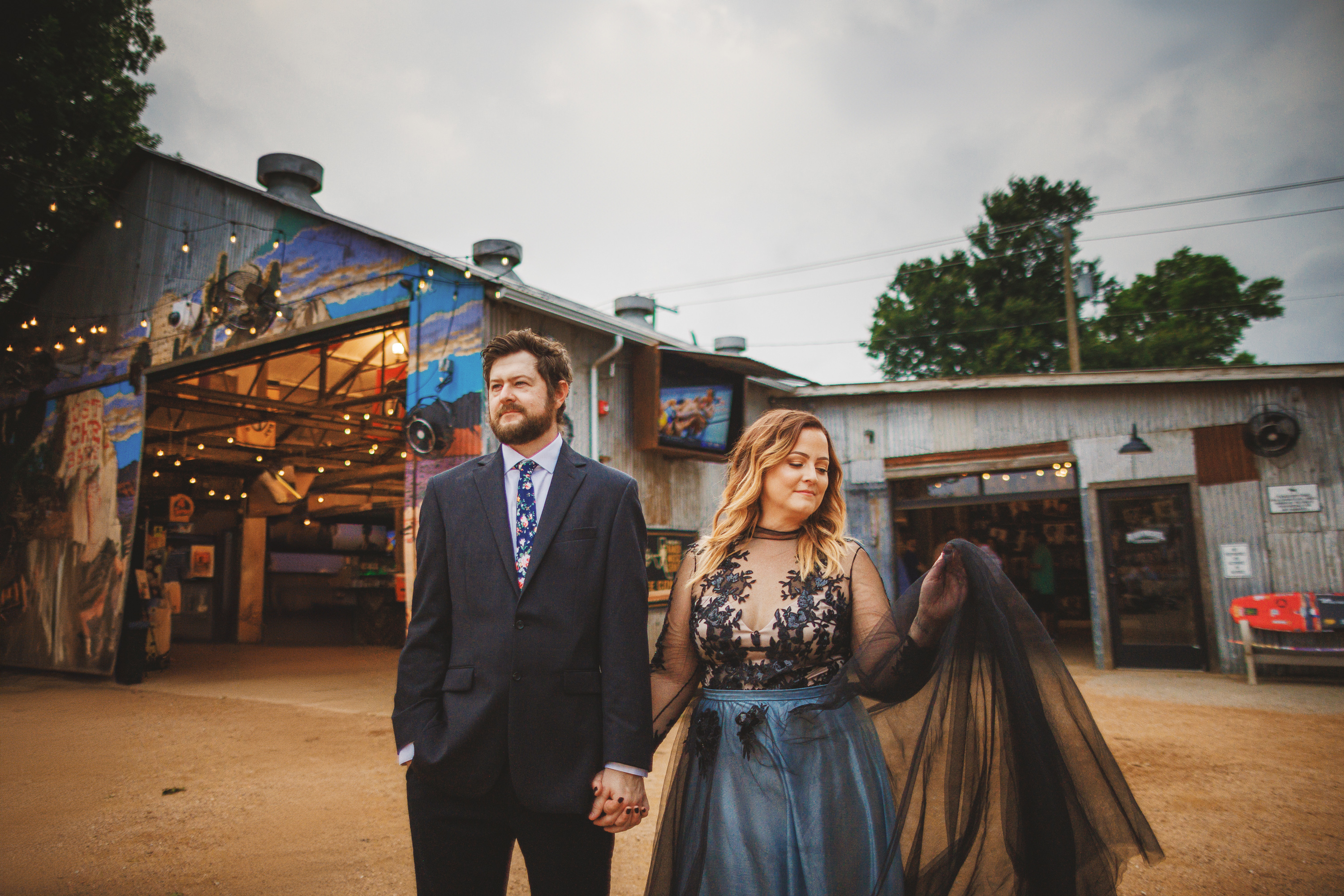 A man in a dark suit and a woman in a dark, semi-sheer dress hold hands outside a rustic, barn-like building adorned with string lights and murals, setting the scene as though composed by an Auto Draft.