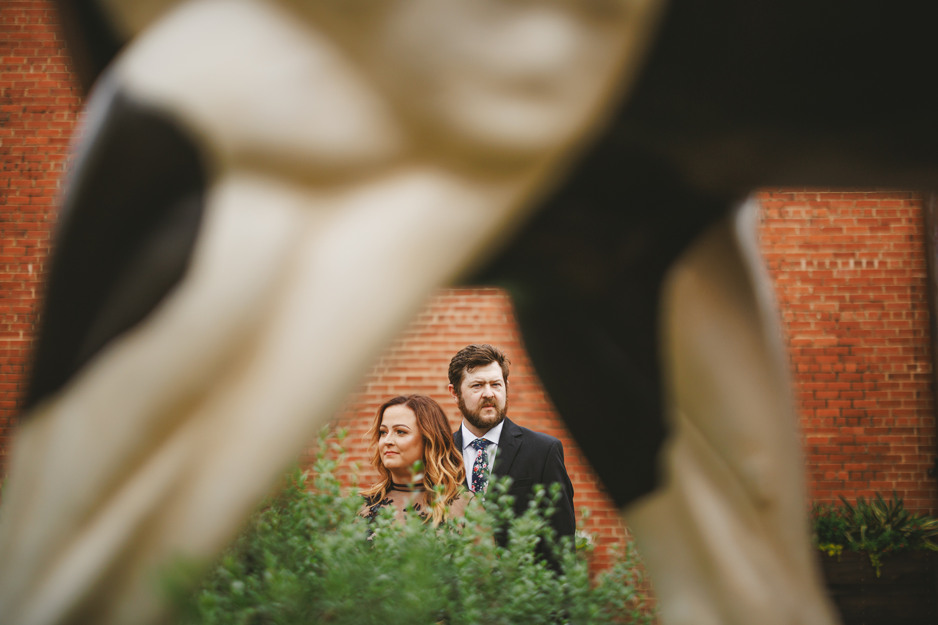 A man and woman are seen through the legs of a statue of a cow, standing in front of a brick building with greenery visible, as if captured in an auto draft.