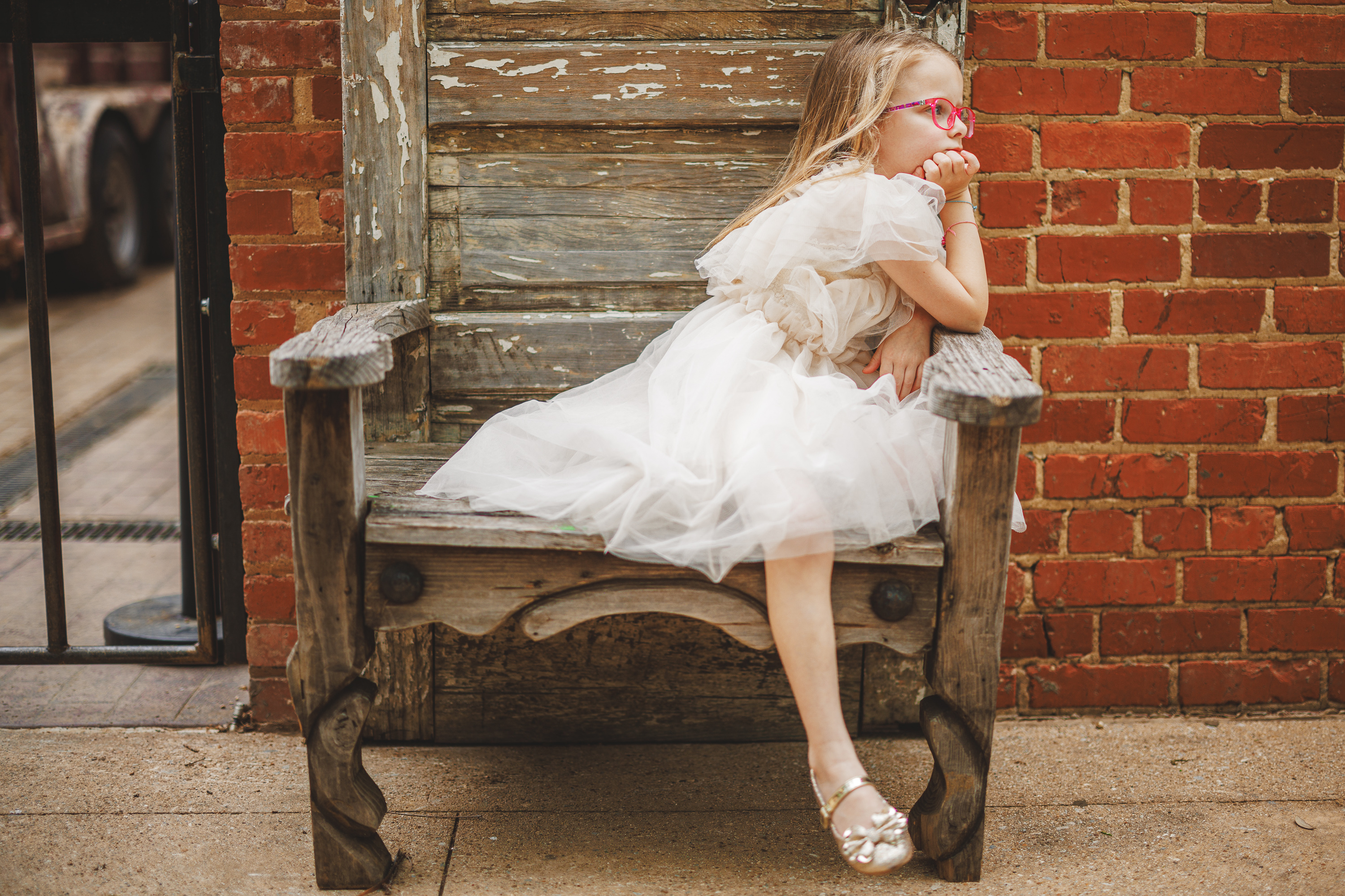 A young girl in a white dress and pink glasses sits on a rustic wooden bench against a brick wall, like an auto draft waiting to be perfected, resting her chin on her hand and looking to the side.
