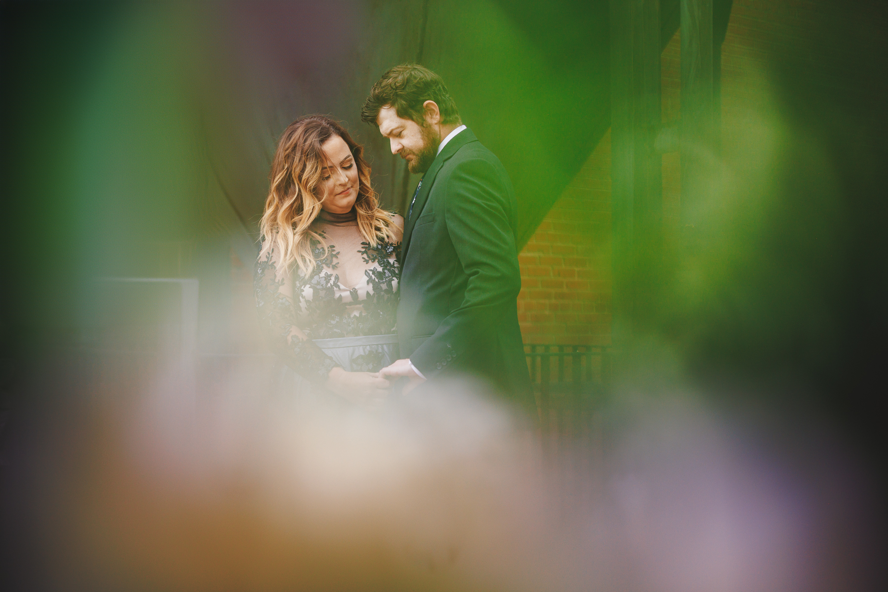 A couple stands close together, the woman in a black dress with lace detail and the man in a dark suit. The image, resembling an auto draft of memories, is partially obscured by blurred, colorful foreground elements.