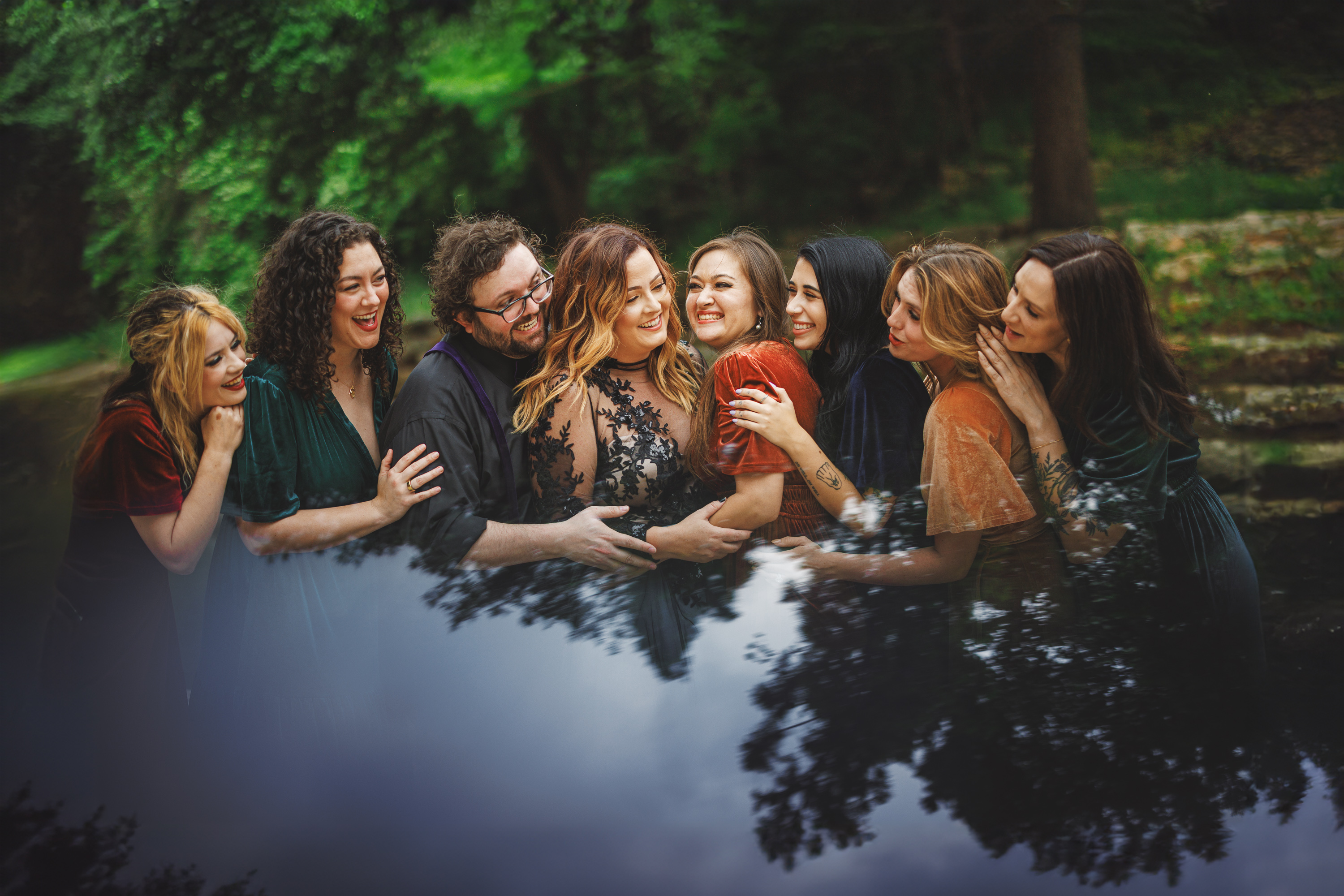 A group of people stands together outdoors, smiling and embracing each other, with greenery in the background and a reflective surface at the bottom of the image. It's almost as if nature itself created this scene on auto draft.