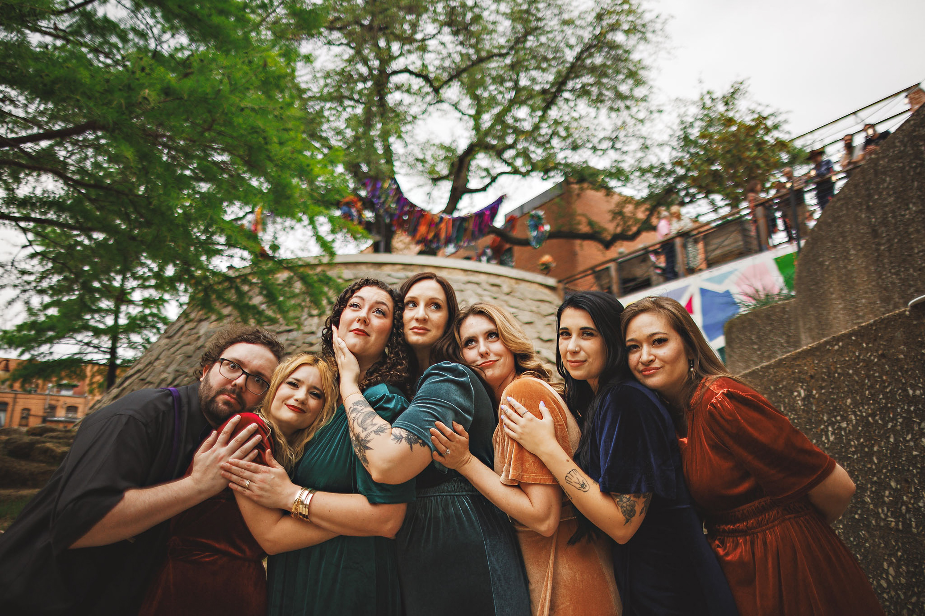 A group of seven people dressed in colorful outfits stand closely together outdoors, posing for a photo. A tree and brick structure are visible in the background, almost like an auto draft scene waiting to be immortalized.