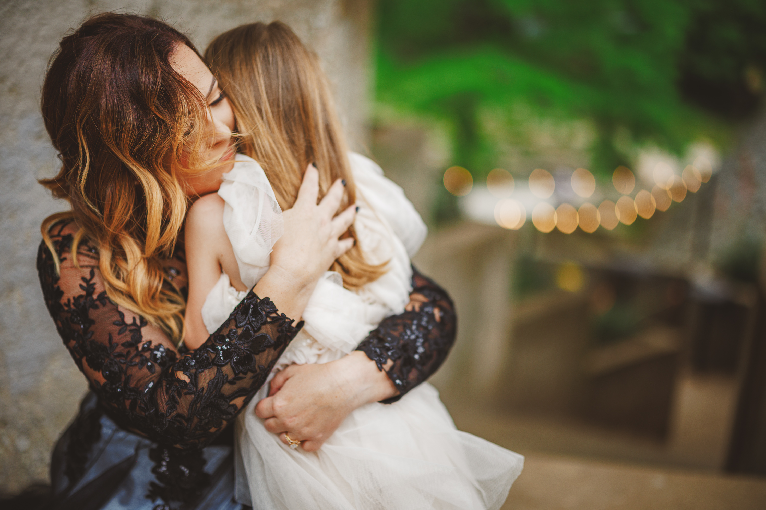 A woman with long, wavy hair hugs a young girl in a white dress on an outdoor staircase. Both have their eyes closed, the background blurred with greenery and string lights, as though captured in an auto draft of cherished memories.