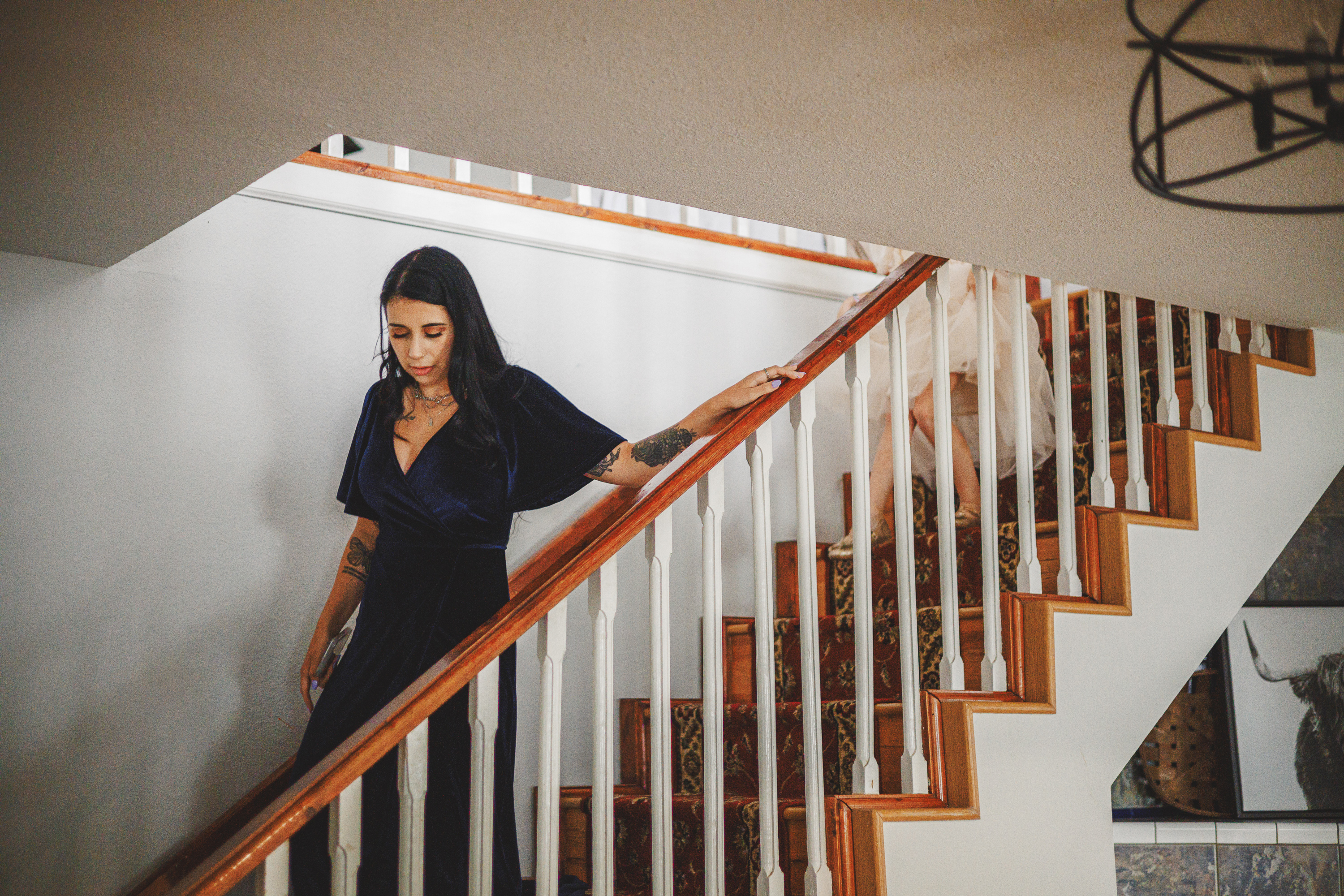 A woman with long dark hair and a navy blue dress, resembling a character from an Auto Draft scene, descends a staircase, holding the handrail. Partial view of another person in the background.