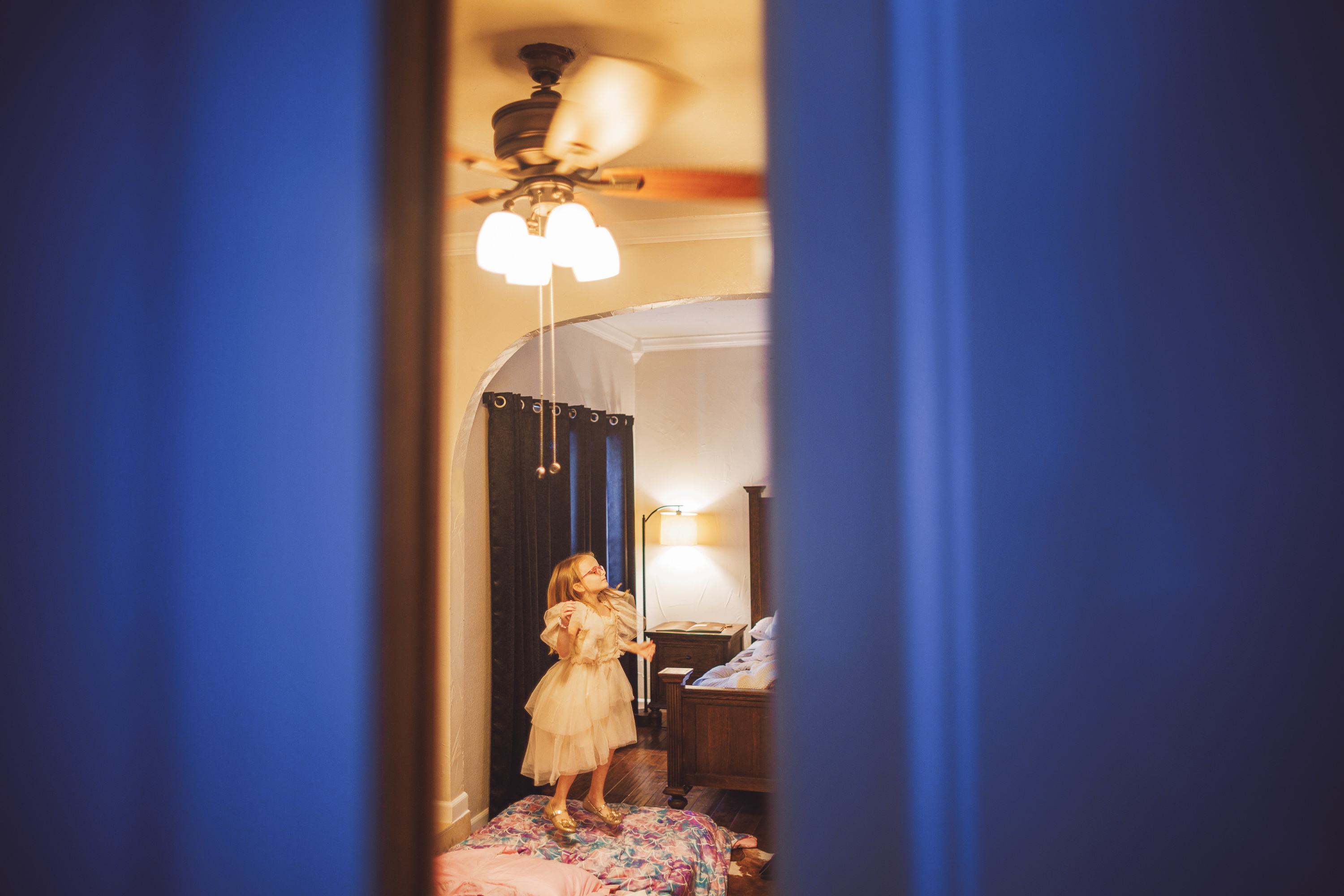 A little girl in a dress stands in a dimly lit room next to a bed, with a ceiling fan and lamp illuminating the scene. The image, caught through an auto draft process, is viewed through a narrow opening, such as a door ajar.