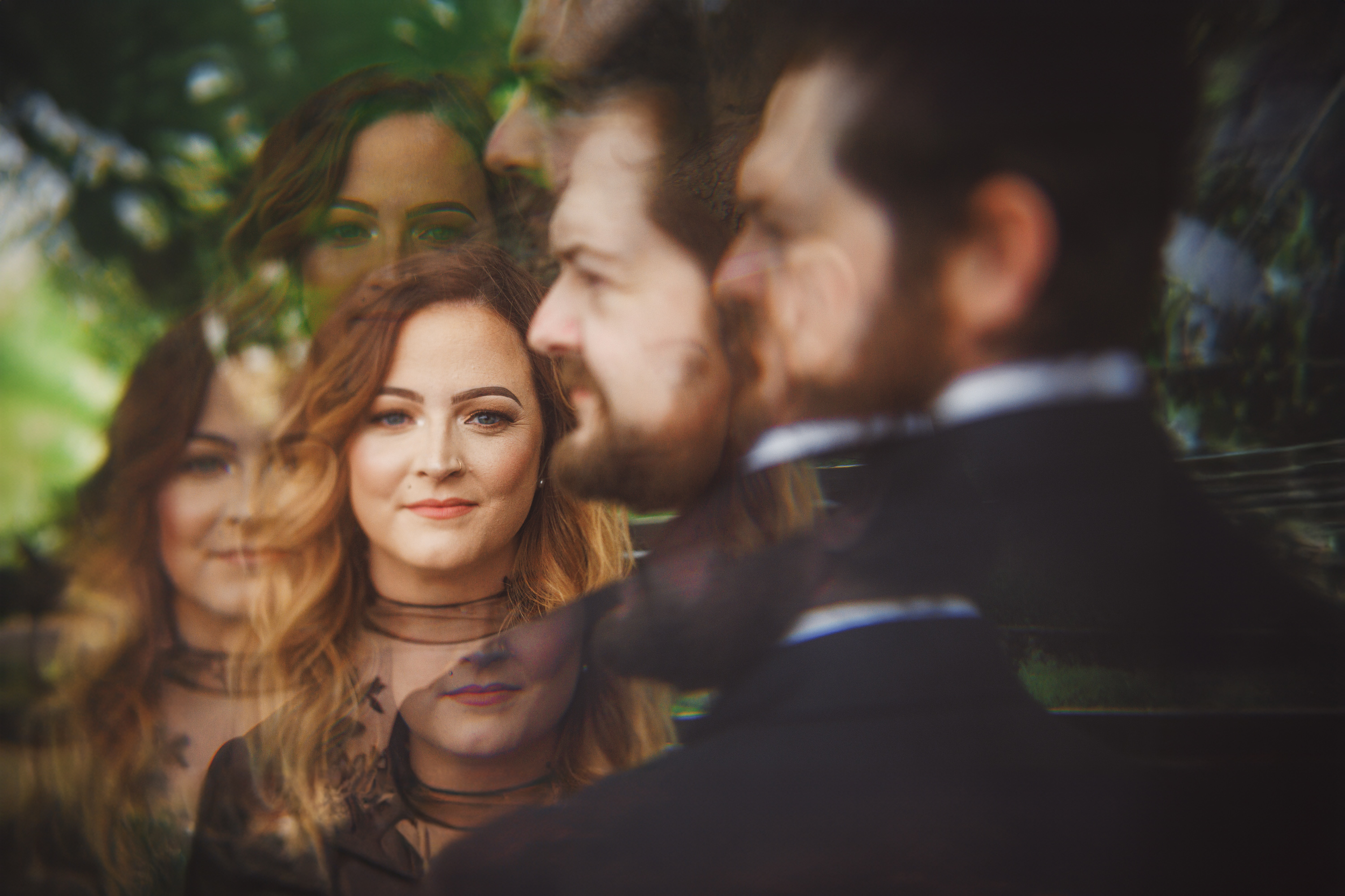 A man and woman with multiple reflections of their faces, reminiscent of an auto draft. The woman has long, wavy hair, and the man has a beard. The background is blurred with green foliage.