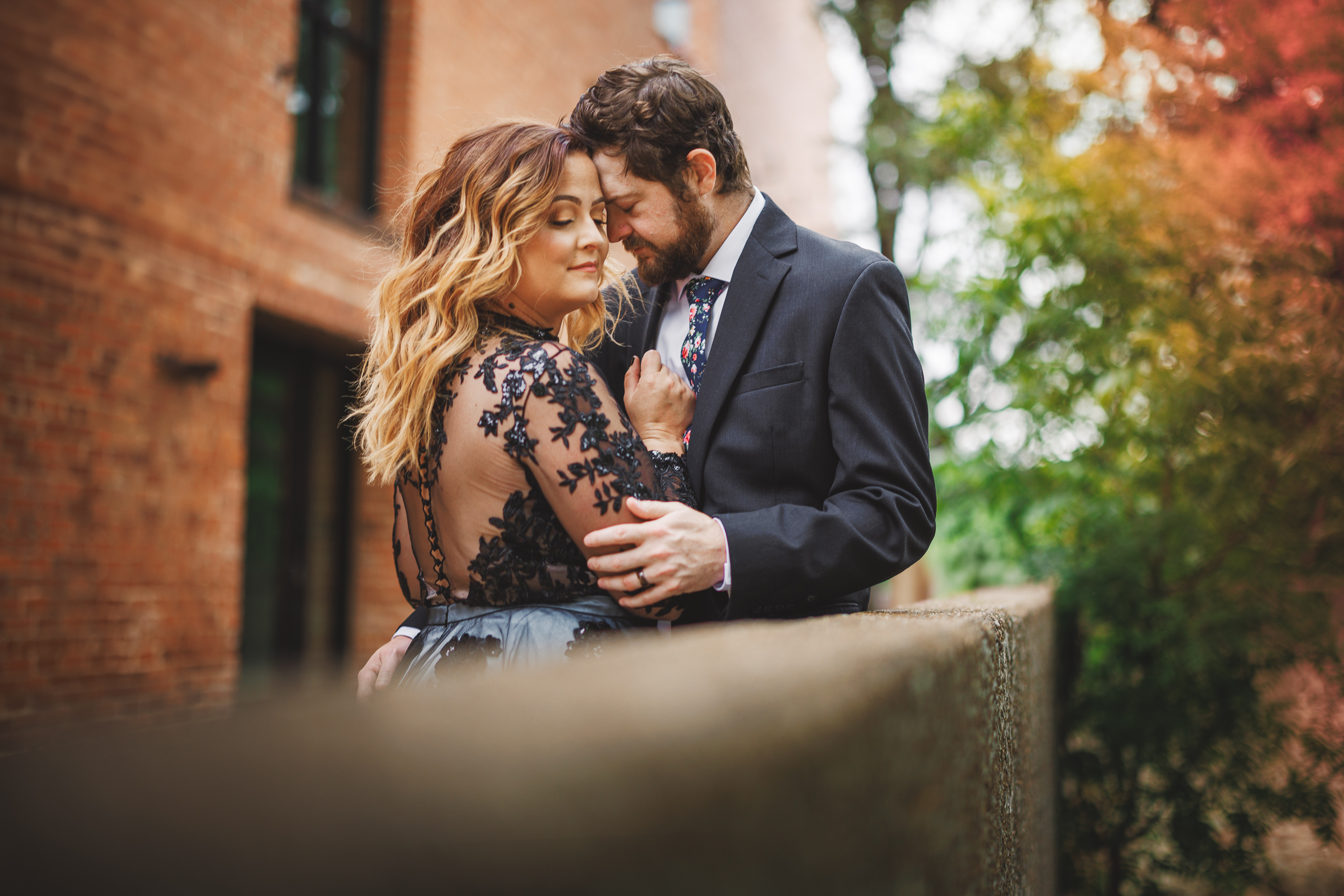 A couple stands close together outdoors near a brick building, with the woman in a black lace dress and the man in a dark suit. They appear affectionate, gently touching foreheads while smiling softly, as if they were the perfect draft of a romantic auto-script.