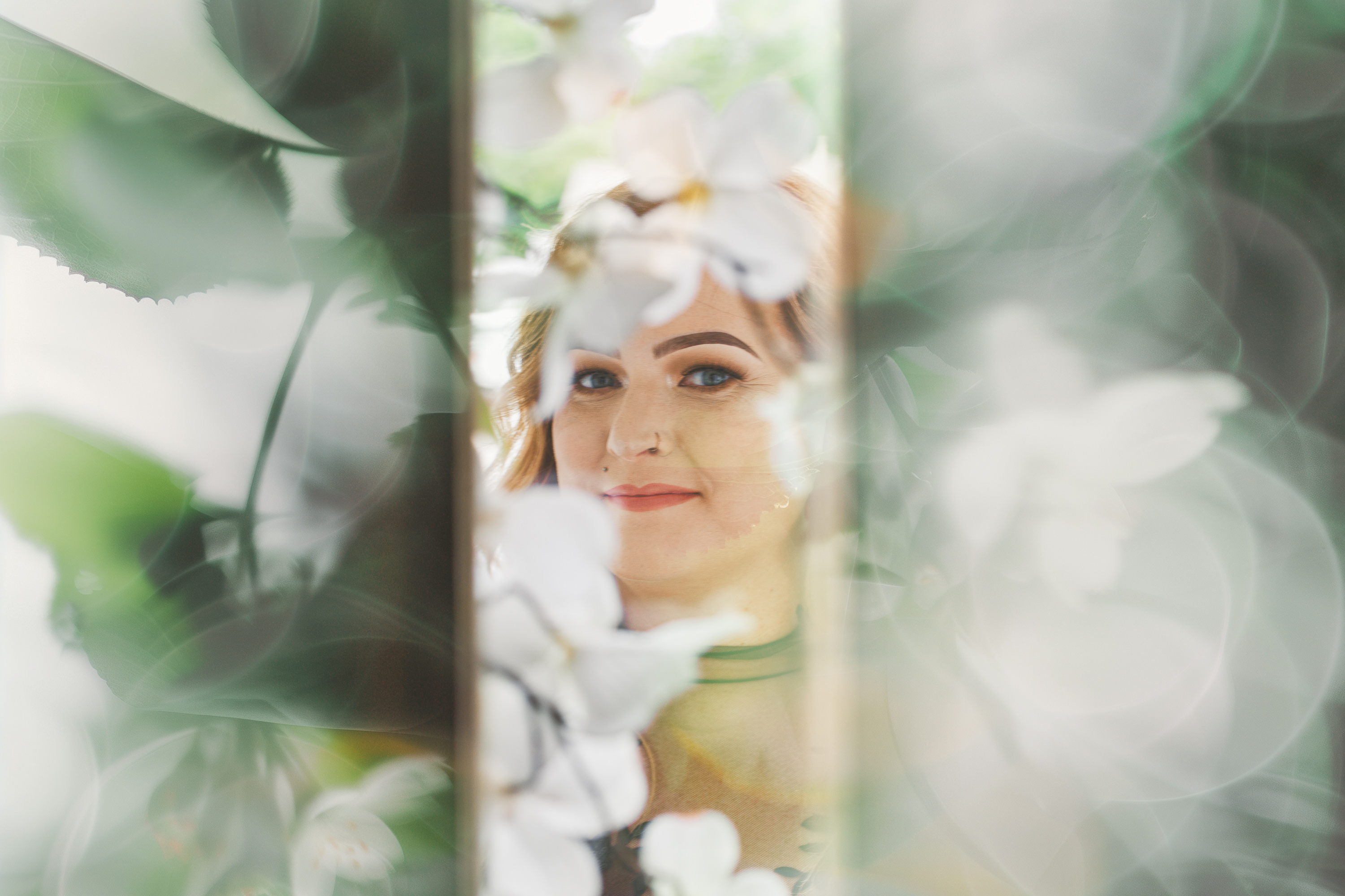 A woman with light hair and makeup is seen through a blurred layer of white flowers and leaves, as if captured in an ethereal Auto Draft painting.