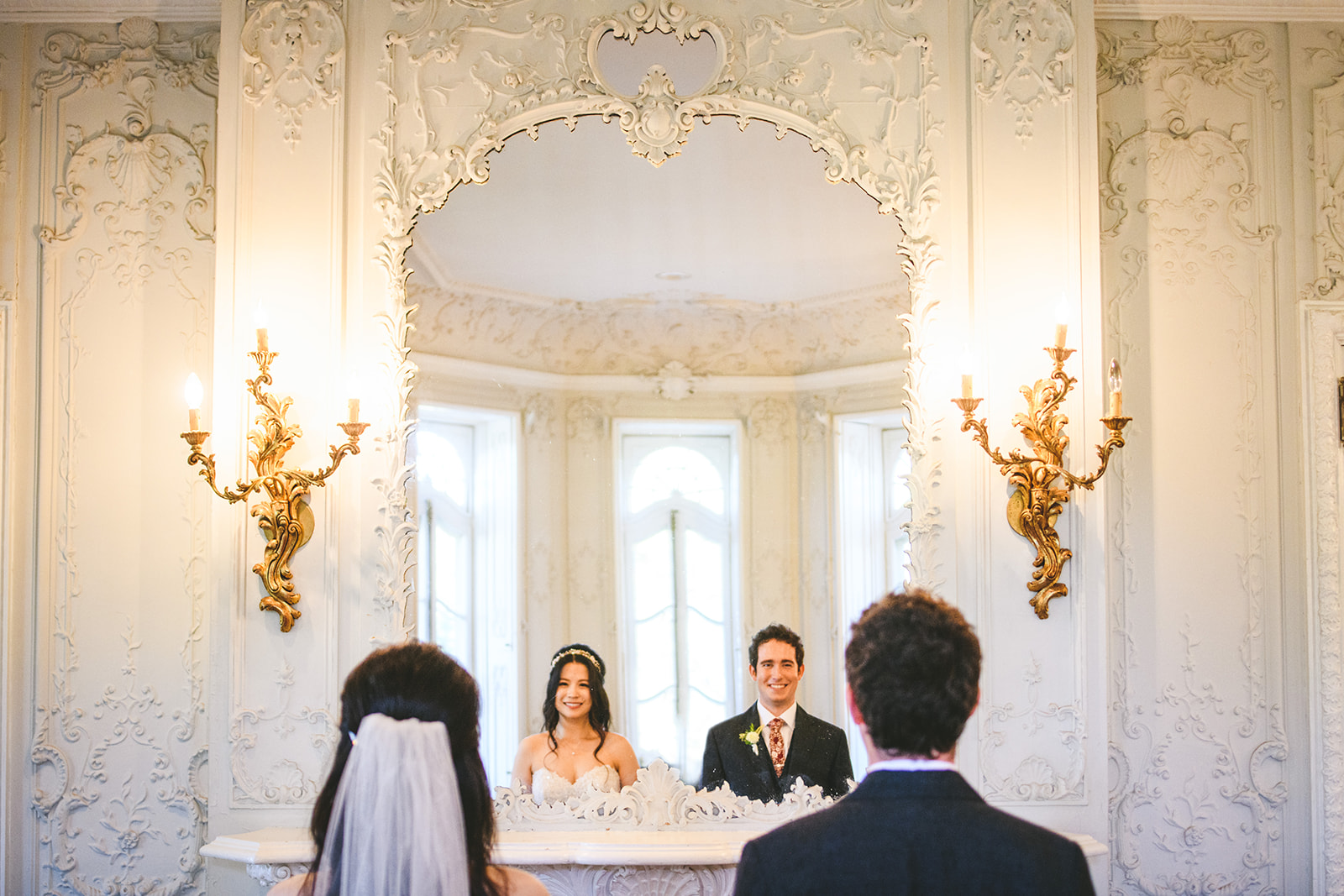 A bride and groom are reflected in a large ornate mirror during a wedding ceremony, their backs to an officiant. The room's decorative wall sconces and intricate moldings add to the elegant ambiance, almost as if every detail was meticulously planned like an auto draft in motion.