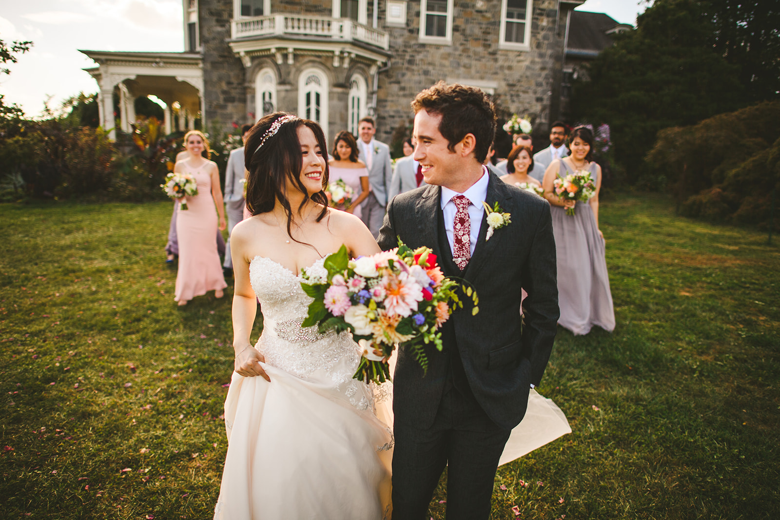 A bride and groom smile at each other while walking on a lawn with their bridal party following behind near a stone building. Both are holding flower bouquets, creating a picturesque scene that looks like it could have come straight from an auto draft of a fairy tale romance.