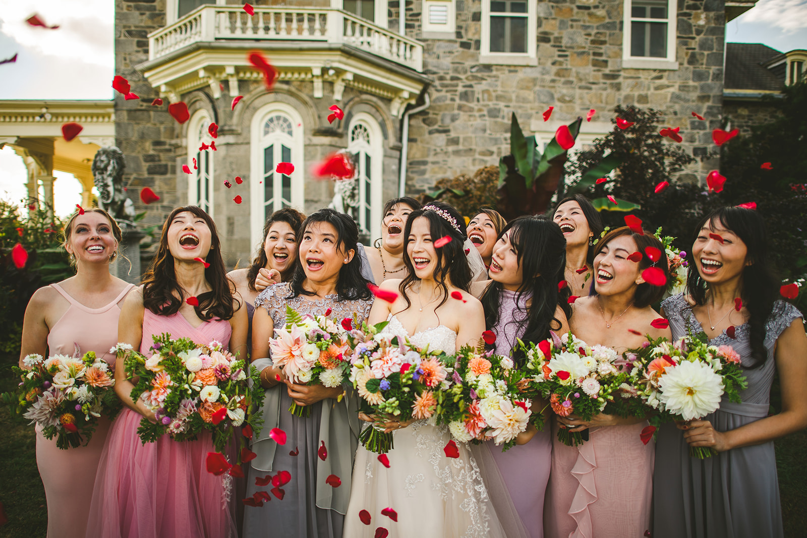 A group of women in formal dresses stand together with bouquets, laughing and smiling, while red rose petals are thrown into the air in front of a large stone building. This picturesque moment feels like it was captured perfectly by an auto draft.