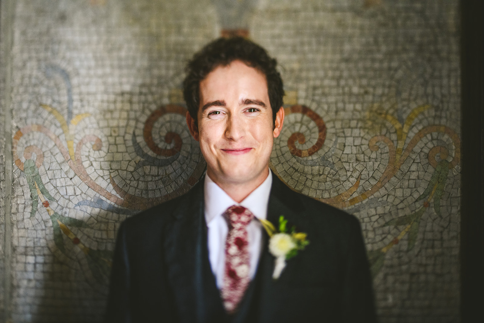 A man in a suit and tie, wearing a white boutonniere, stands in front of a decorative mosaic wall, smiling at the camera, as if ready for an Auto Draft event.