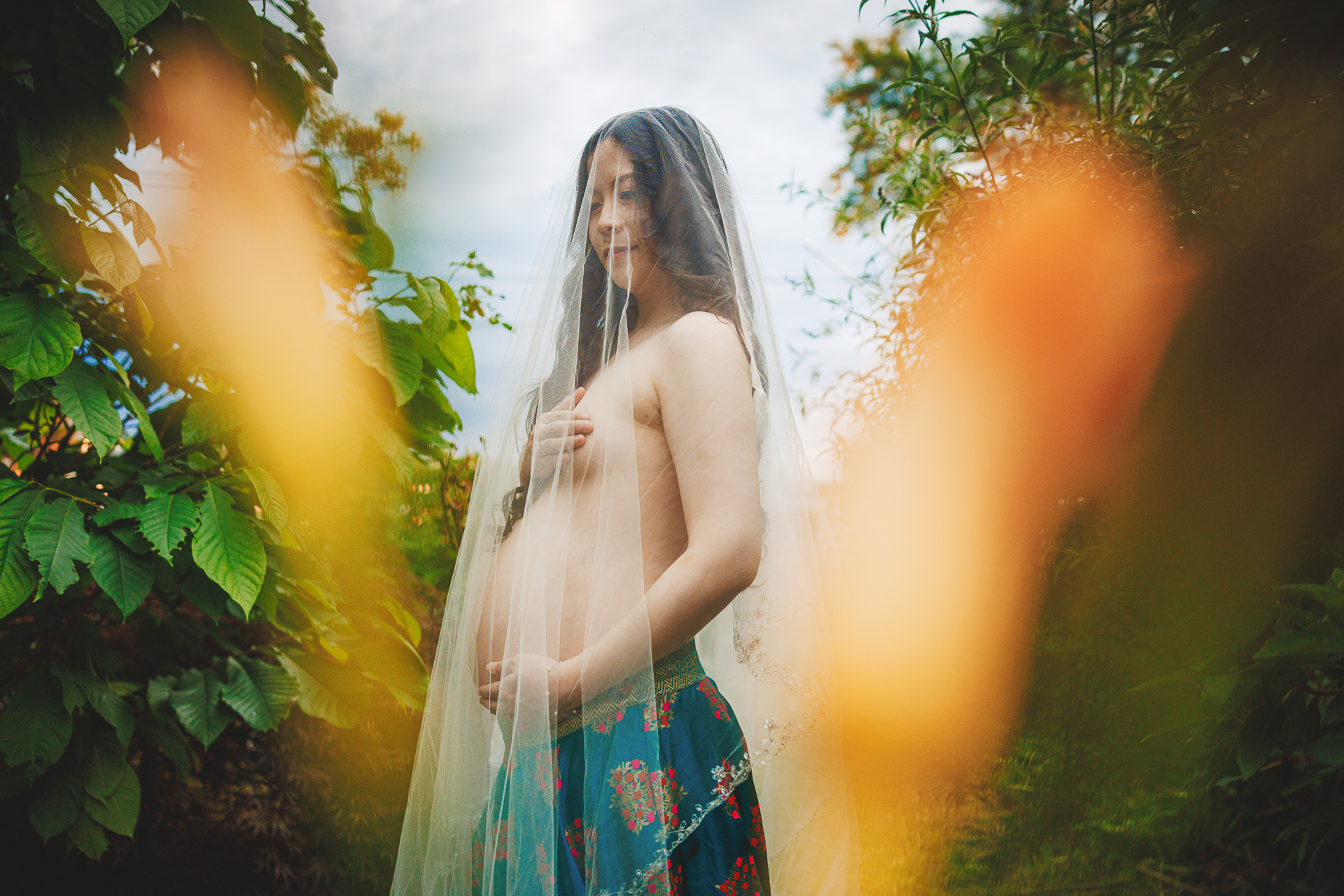A pregnant person, partially covered by a delicate veil, stands outdoors with an enchanting background of foliage and flowers, as if nature itself provided an auto draft for this serene moment.