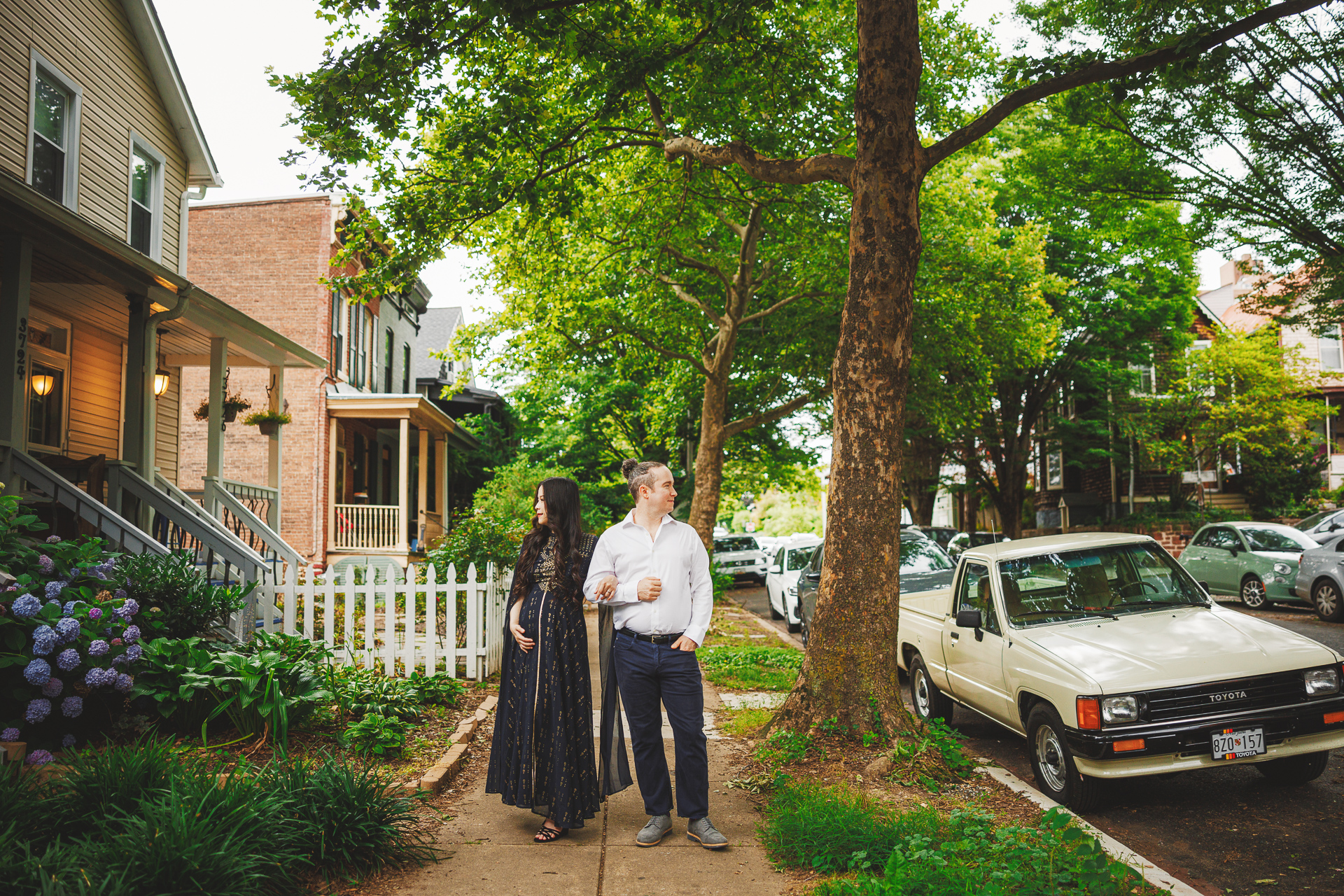 A man and a woman walk along a tree-lined street with houses and parked cars. The woman holds a coffee cup as the man has his hands in his pockets, their steps an auto draft of familiarity and ease.