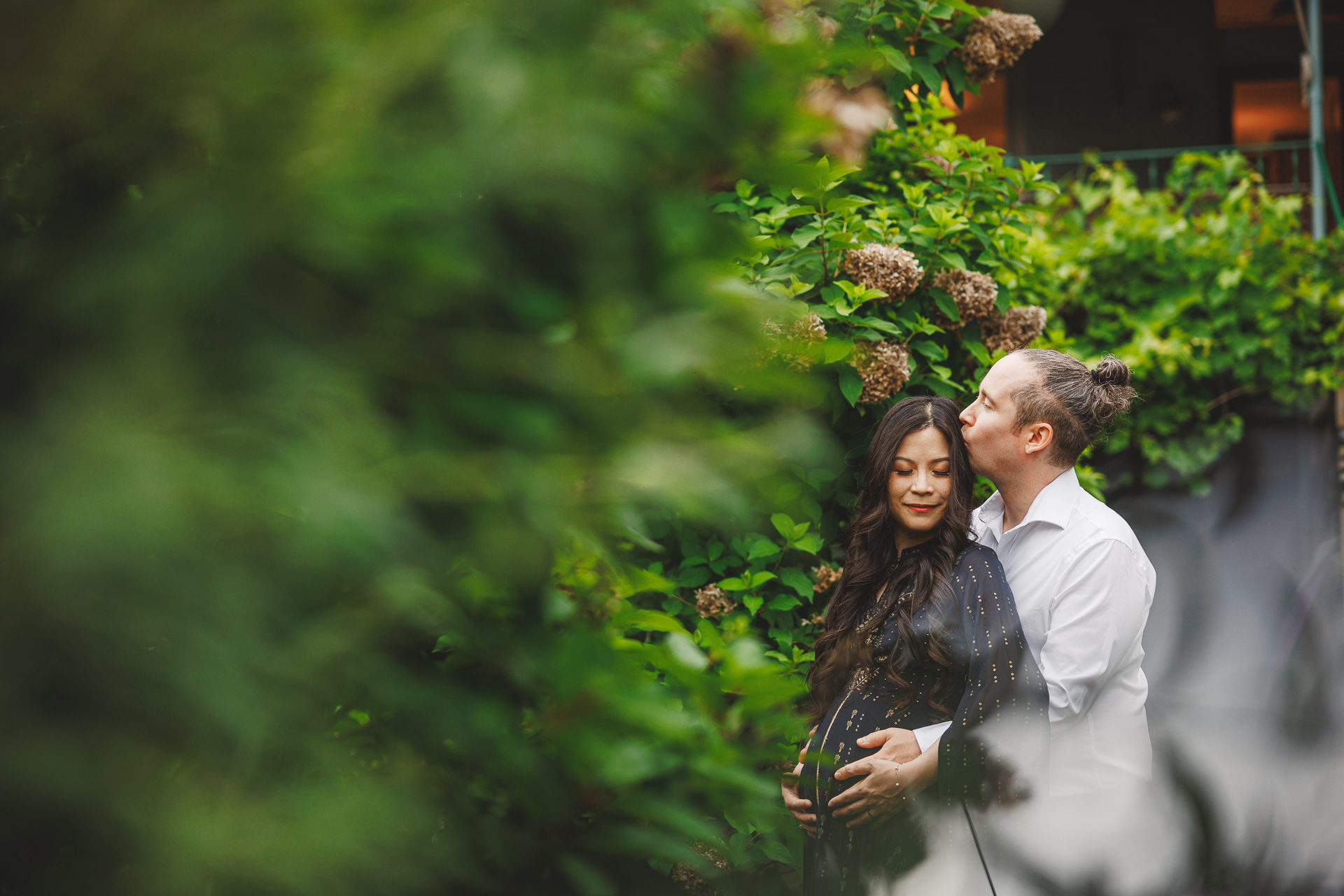 A man stands behind a pregnant woman, with his arms around her and kissing her head. Amid the lush greenery and flowers, it feels like a moment auto-drafted by nature itself.