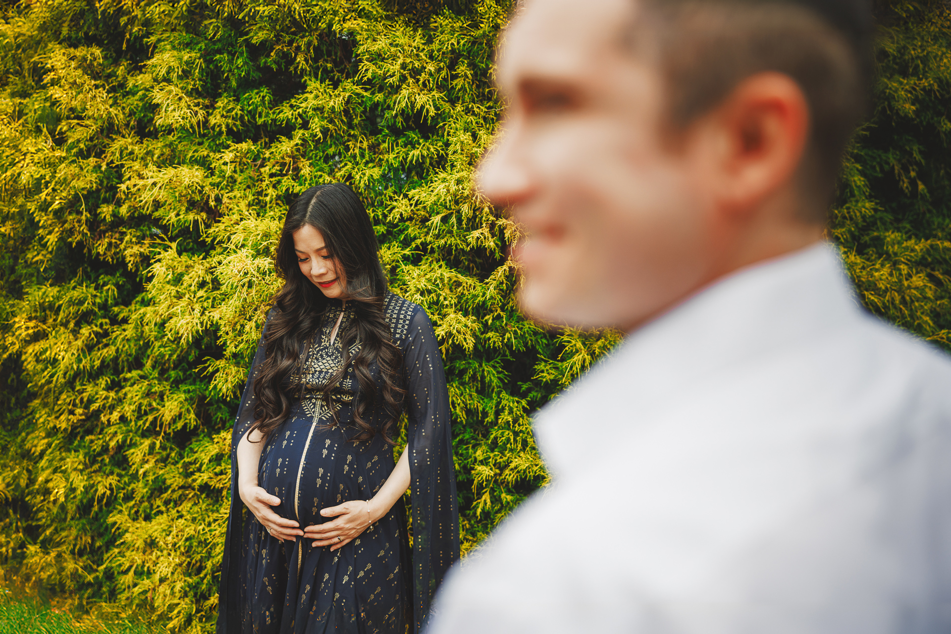 A pregnant woman in a long dress holds her belly and looks down, as if deep in thought. A man, blurred in the foreground like an auto draft, stands nearby. They are surrounded by lush greenery.
