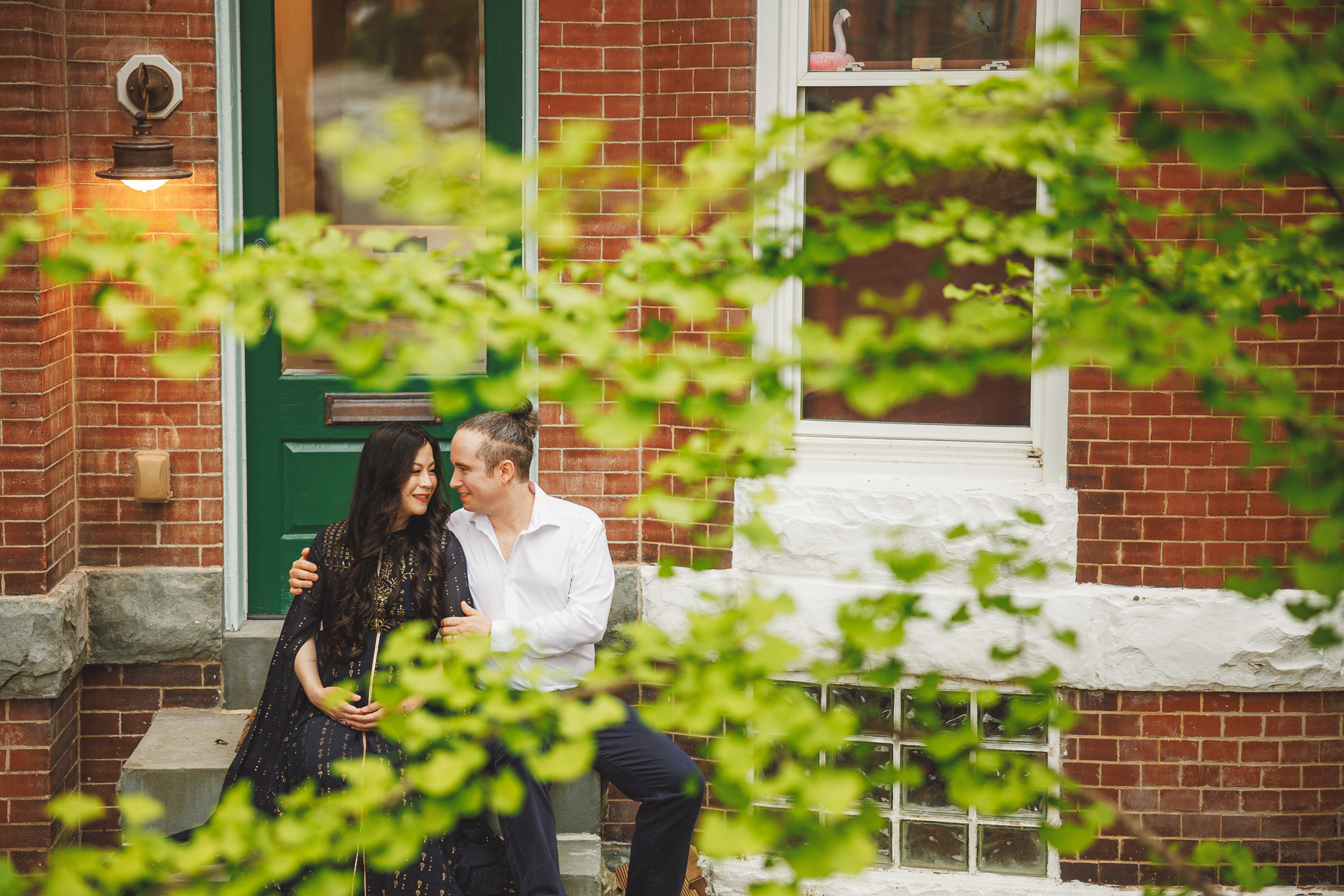 A couple sits close together on the steps of a brick building with a green door, partially obscured by leafy branches, as if straight from an auto draft of a romantic scene.