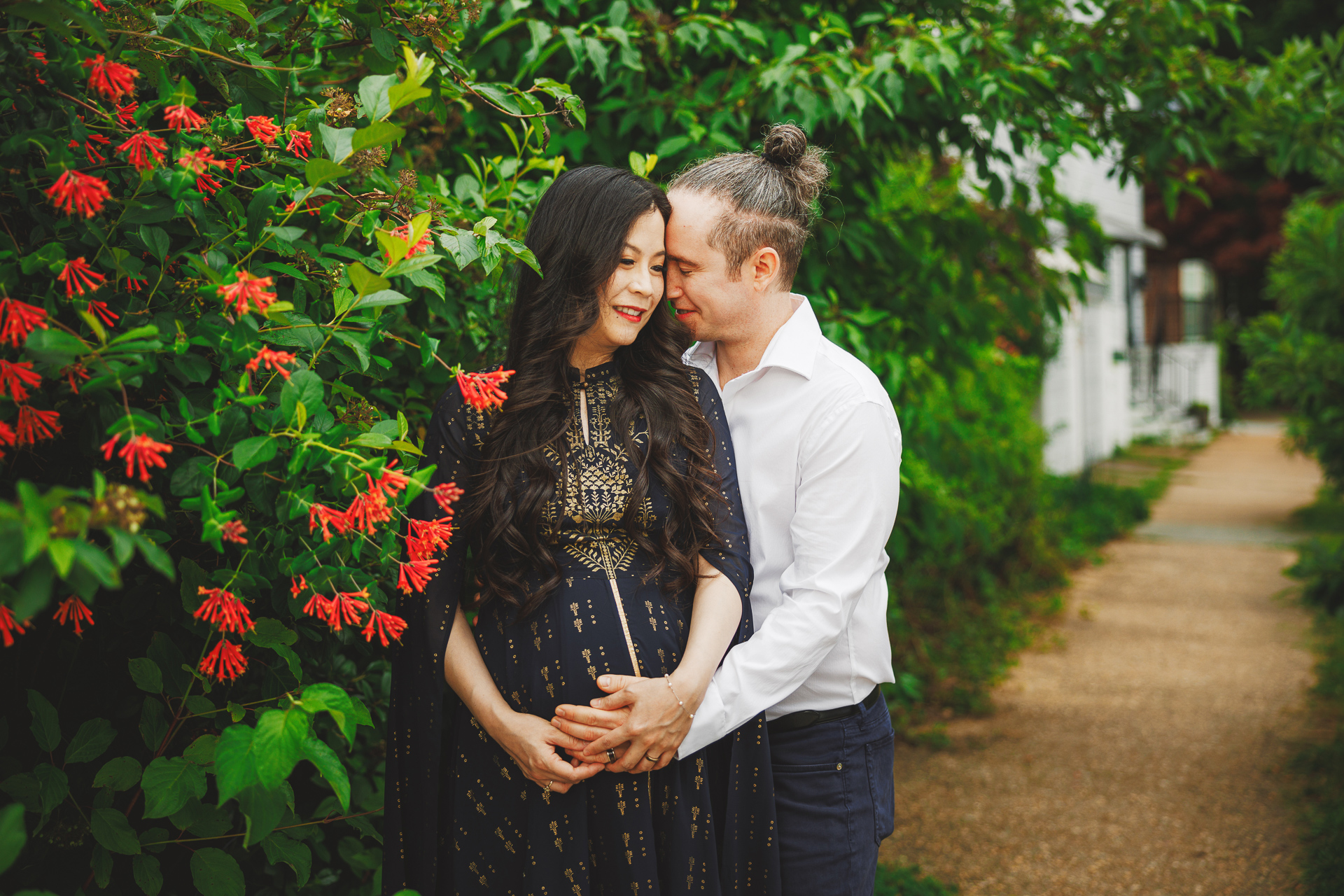 A man in a white shirt embraces a woman in a black embroidered dress surrounded by greenery and red flowers on the pathway, creating an image that feels like an auto draft of pure romance.