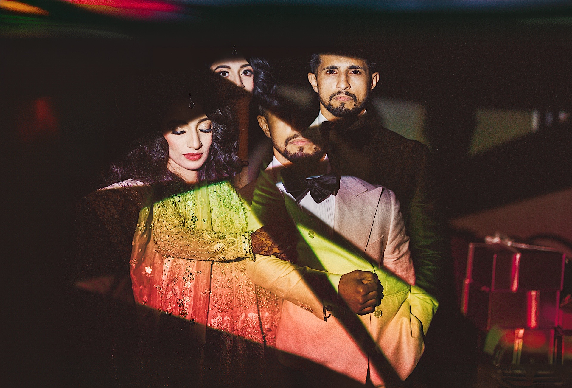 A couple stands close together, with colorful light projections creating abstract patterns on their faces and clothing in a dimly lit setting, resembling an Auto Draft of a modern art piece.