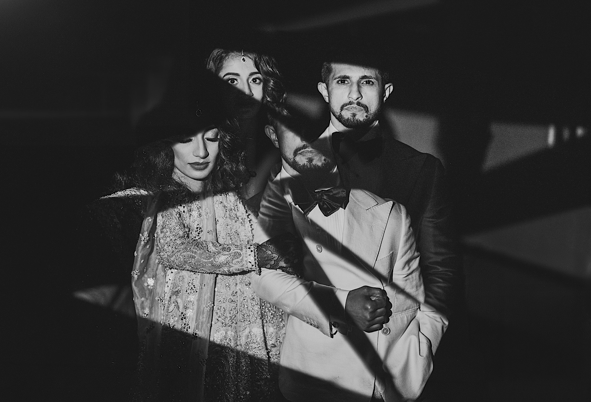 Black and white image of four people dressed in formal attire, with dramatic lighting casting shadows across their faces, creating an almost auto draft-like contrast.