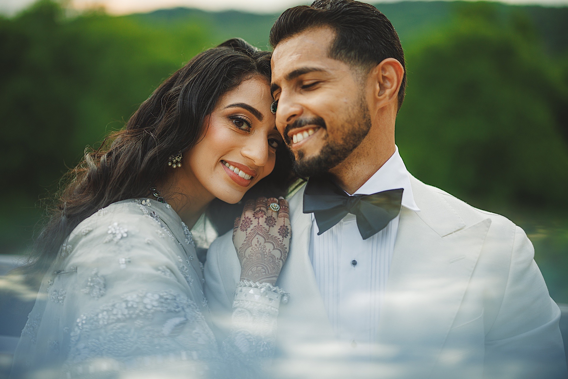 A smiling couple dressed in formal attire, embracing with nature in the background. The woman's hand, adorned with intricate henna patterns, gently touches the man's chin while they stand as if time itself were on auto draft, capturing their serene moment perfectly.