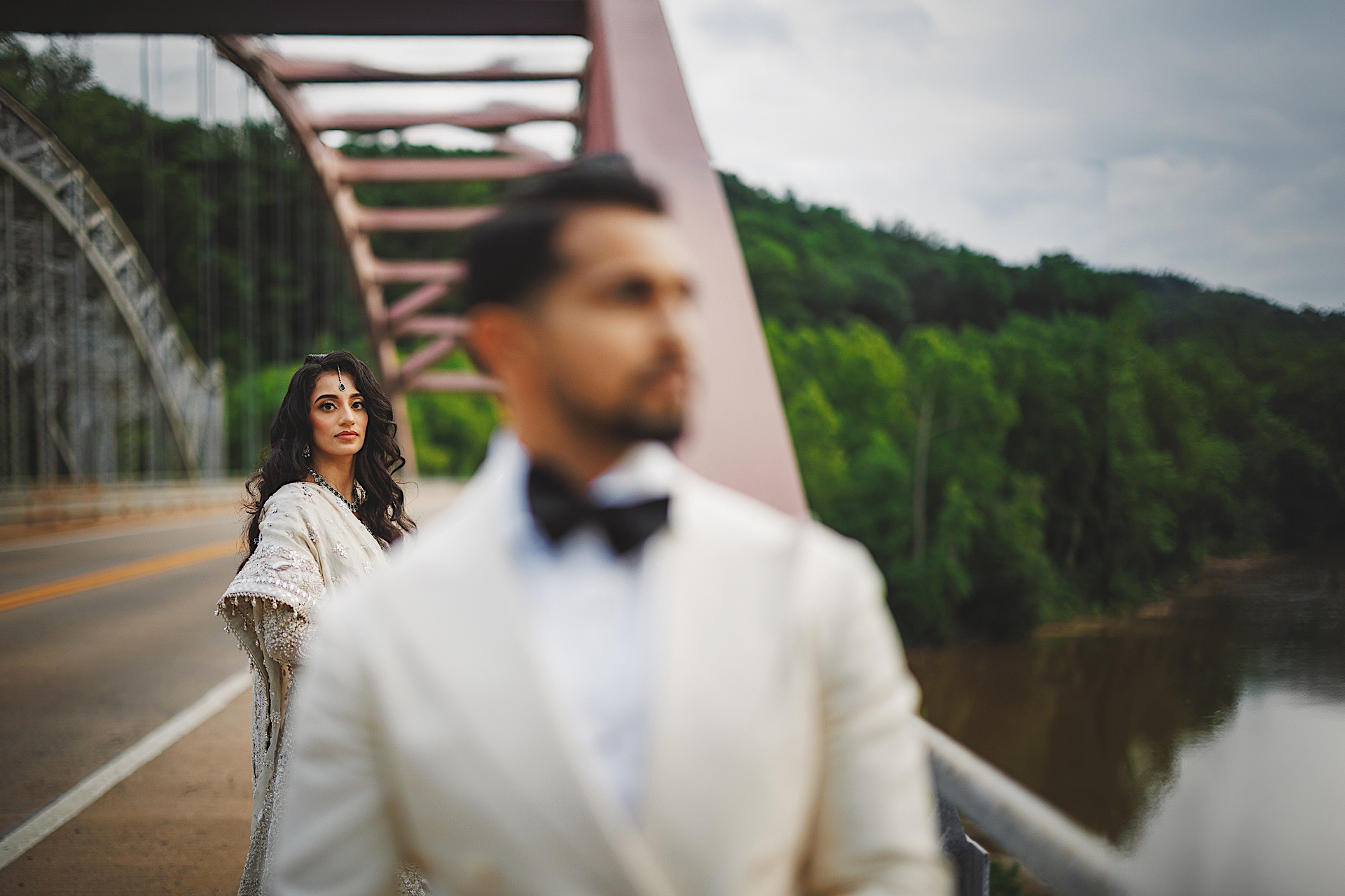 A woman in traditional attire stands on a bridge, looking into the distance, with a man in a suit and bow tie in the foreground, slightly out of focus. Lush greenery and a river stretch out behind them, almost like an auto draft of a painter's dream.