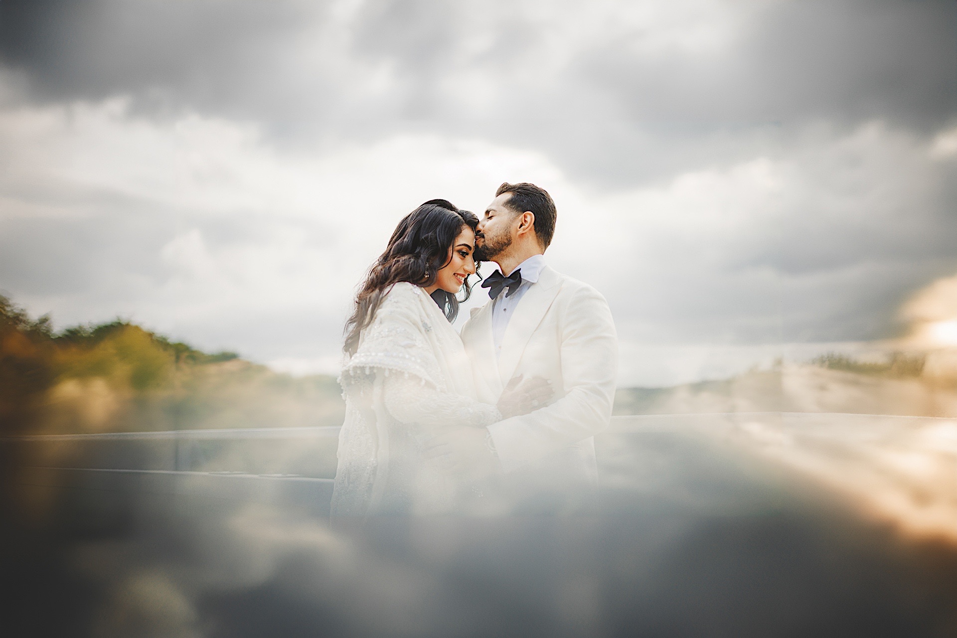 A couple stands together outdoors, the man gently kissing the woman's forehead. They are dressed in formal attire under a cloudy sky, creating a scene that feels like it’s been lifted straight out of an auto-drafted romance novel.