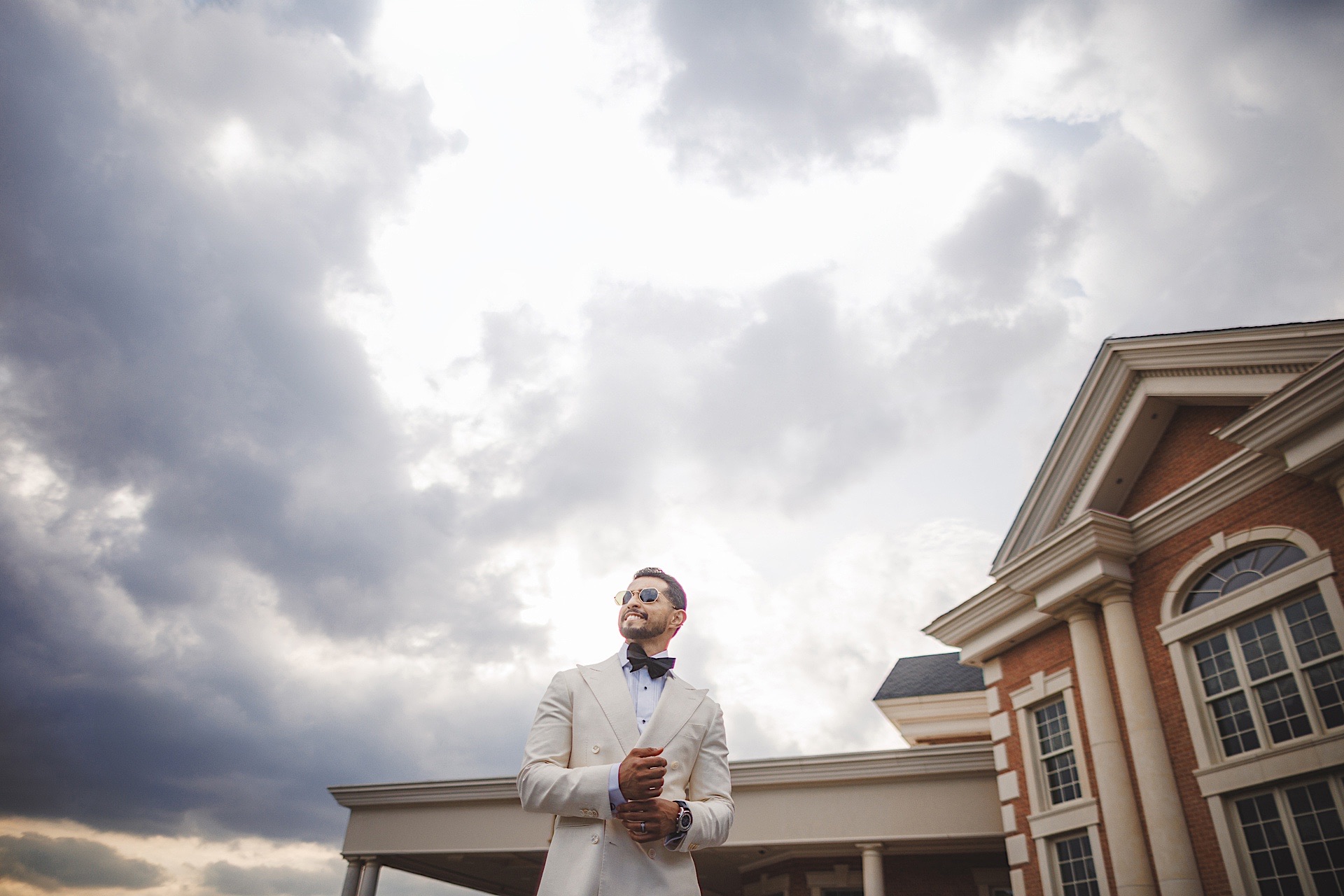A man in a light suit and bow tie stands outside a large building with columns and arched windows, under a partly cloudy sky, reminiscent of an Auto Draft sketch taking shape.