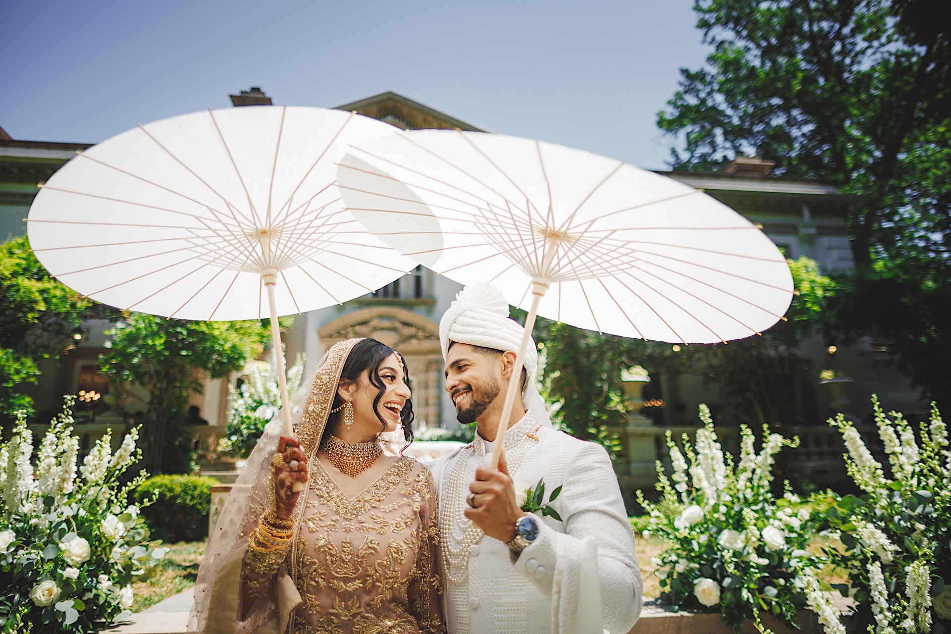 A bride and groom in traditional attire stand outdoors, smiling at each other while holding white parasols. The background features lush greenery and the elegant facade of a large building, creating a picturesque scene that looks like it could be an Auto Draft of a timeless romance.