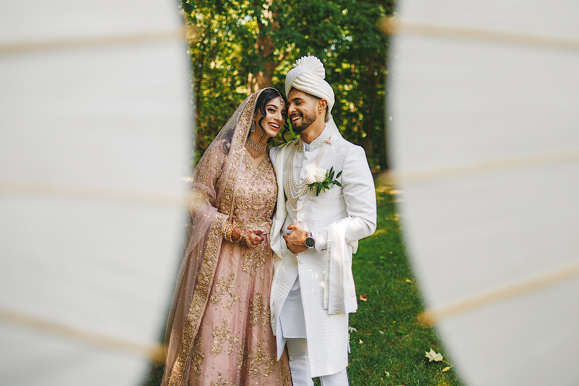 A bride and groom in traditional attire stand closely together, smiling, with a green, leafy background seen through a curtain-like frame, almost as if crafted by an artistic auto draft.