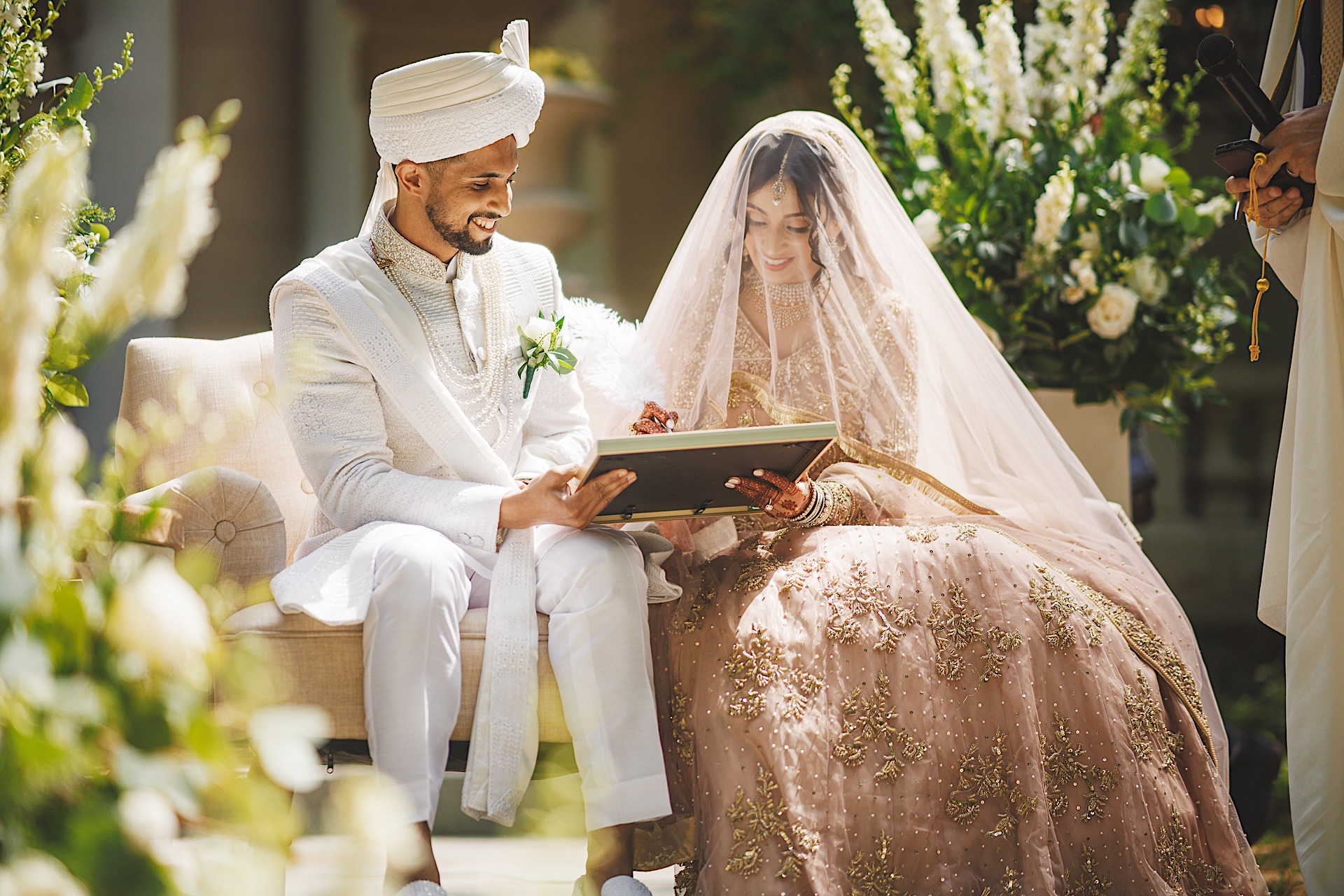 A bride and groom, dressed in elegant traditional attire, sit together during their wedding ceremony. The bride holds a framed document titled "Auto Draft," both looking at it with smiles.
