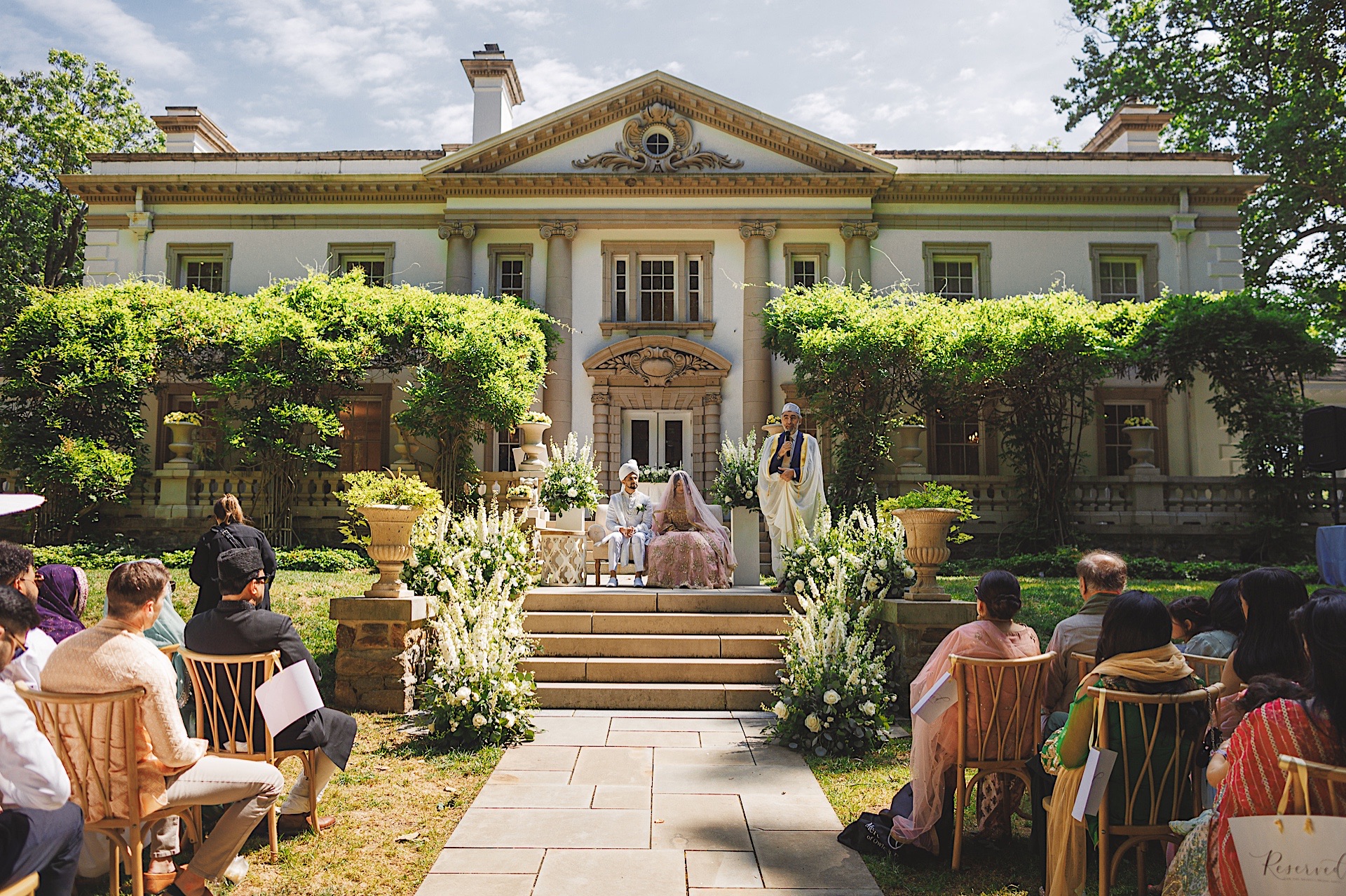 A wedding ceremony taking place in a garden in front of a large, ornate building. Guests are seated on either side of an aisle lined with flowers, as if orchestrated by an impeccable Auto Draft. The couple and officiant are positioned on a decorated stage.
