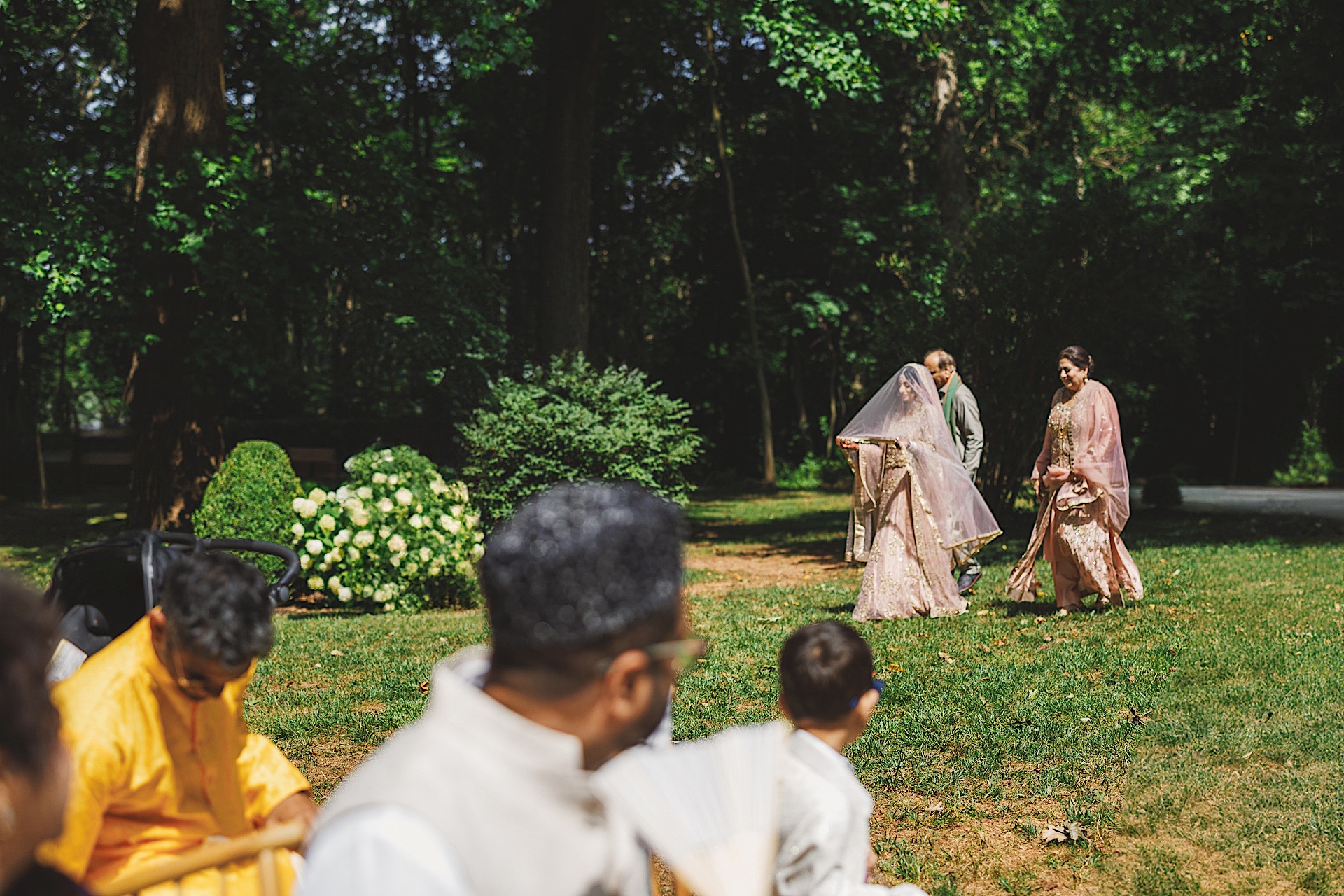 A bride, covered by a sheer veil and wearing traditional attire, walks in a garden, accompanied by an older woman. Several seated guests in colorful clothing observe the scene like an auto draft of vibrant moments. The background is lush and green.