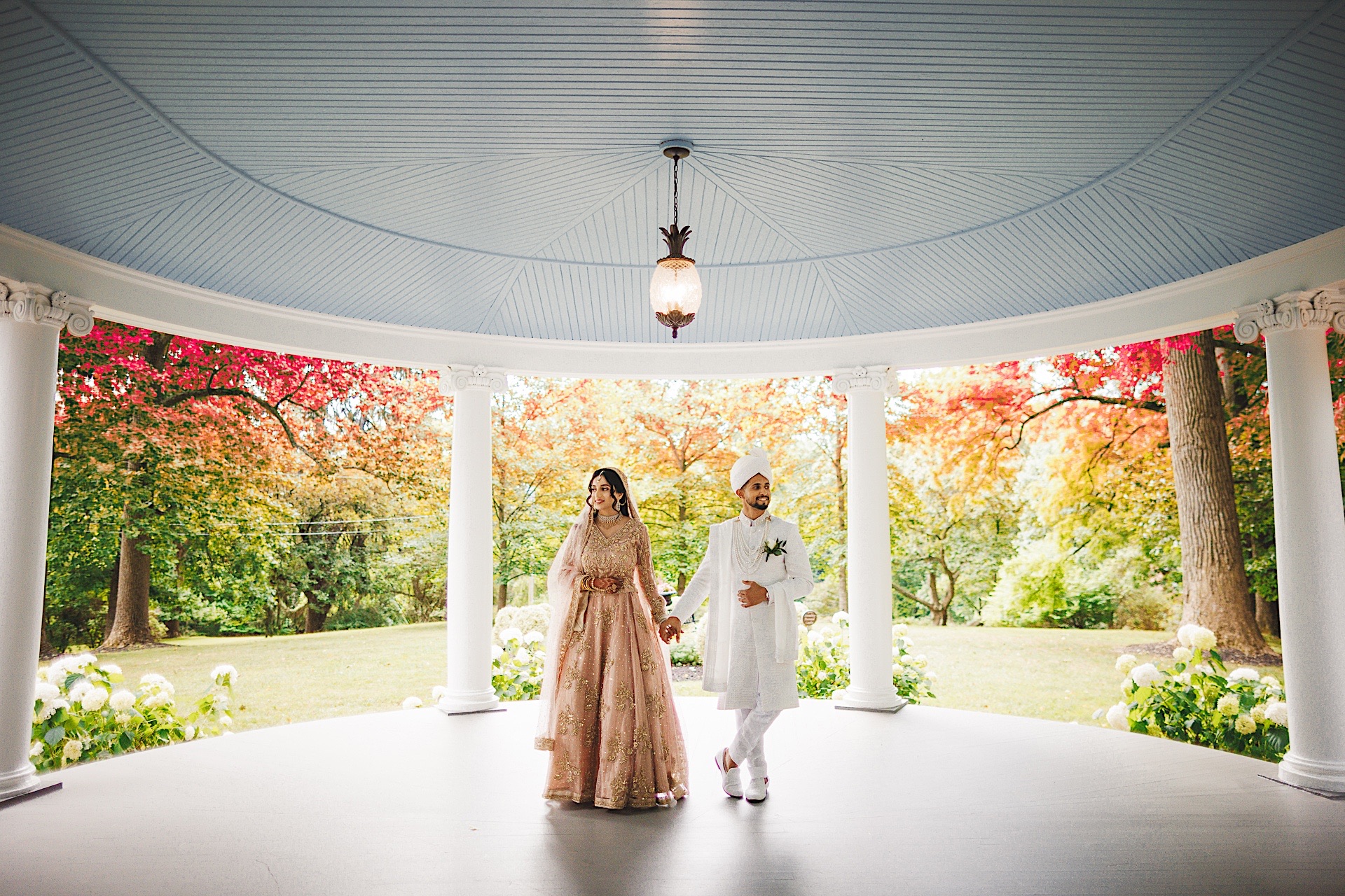 A couple dressed in traditional attire stands in a gazebo, surrounded by vibrant autumn foliage, reminiscent of an auto draft sketch brought to life.