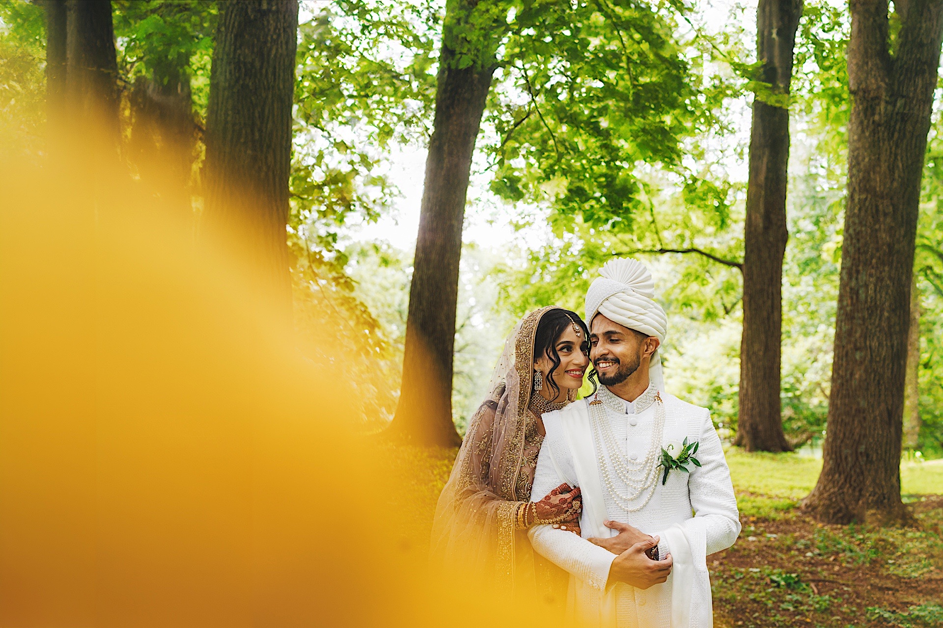 A couple in traditional attire stands closely together under tall trees, smiling and embracing. The background is lush and green, with a yellow-orange blur partially obscuring the foreground, as if capturing an auto draft of nature's splendor.