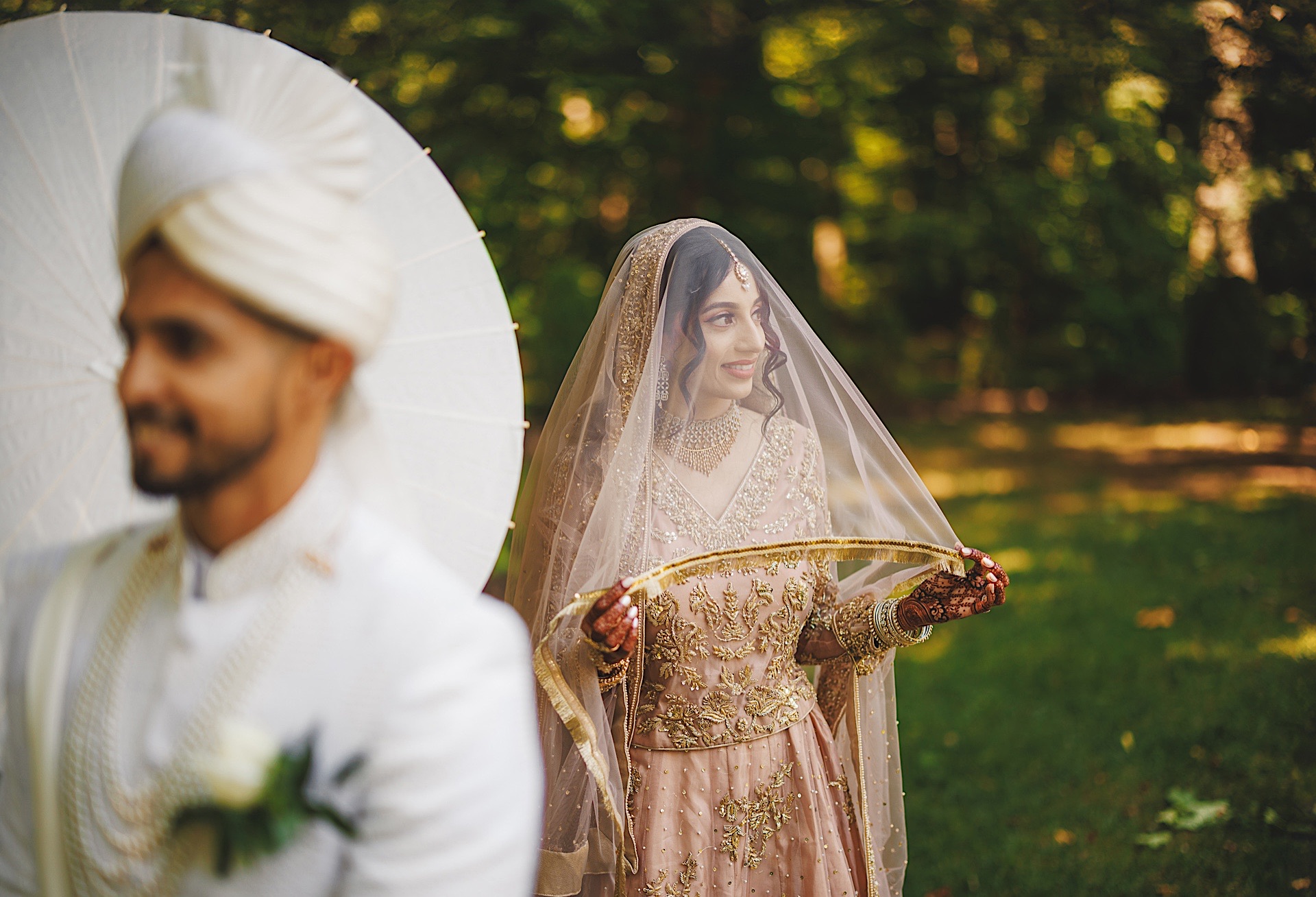 A South Asian bride in a gold and pink outfit smiles behind her groom in white with a parasol, captured beautifully by a wedding photographer under the bright Maryland sun.