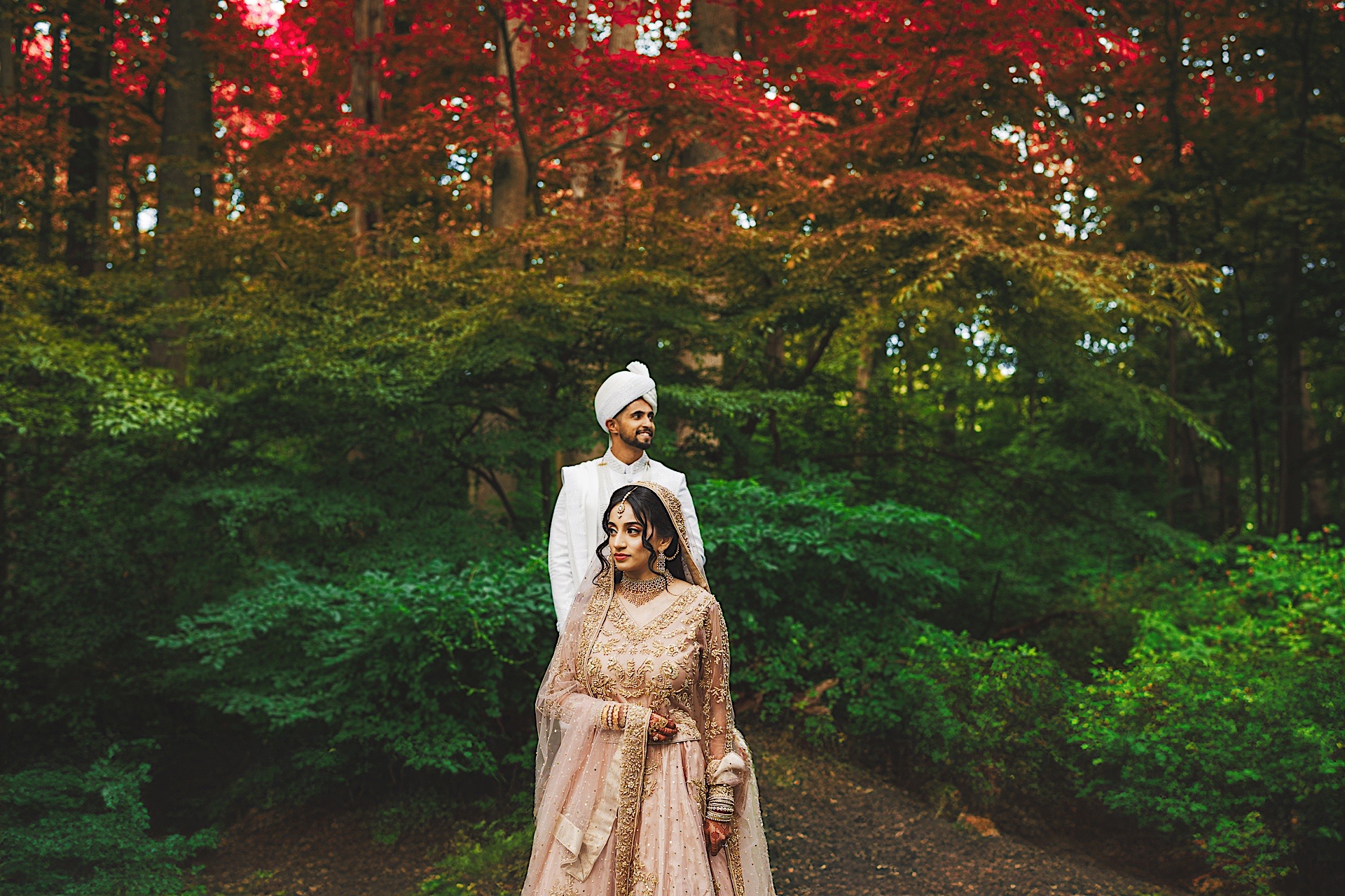 A couple dressed in traditional attire stands outdoors against a backdrop of lush greenery and vibrant red foliage, capturing a moment that seems almost like an auto draft of nature's beauty.