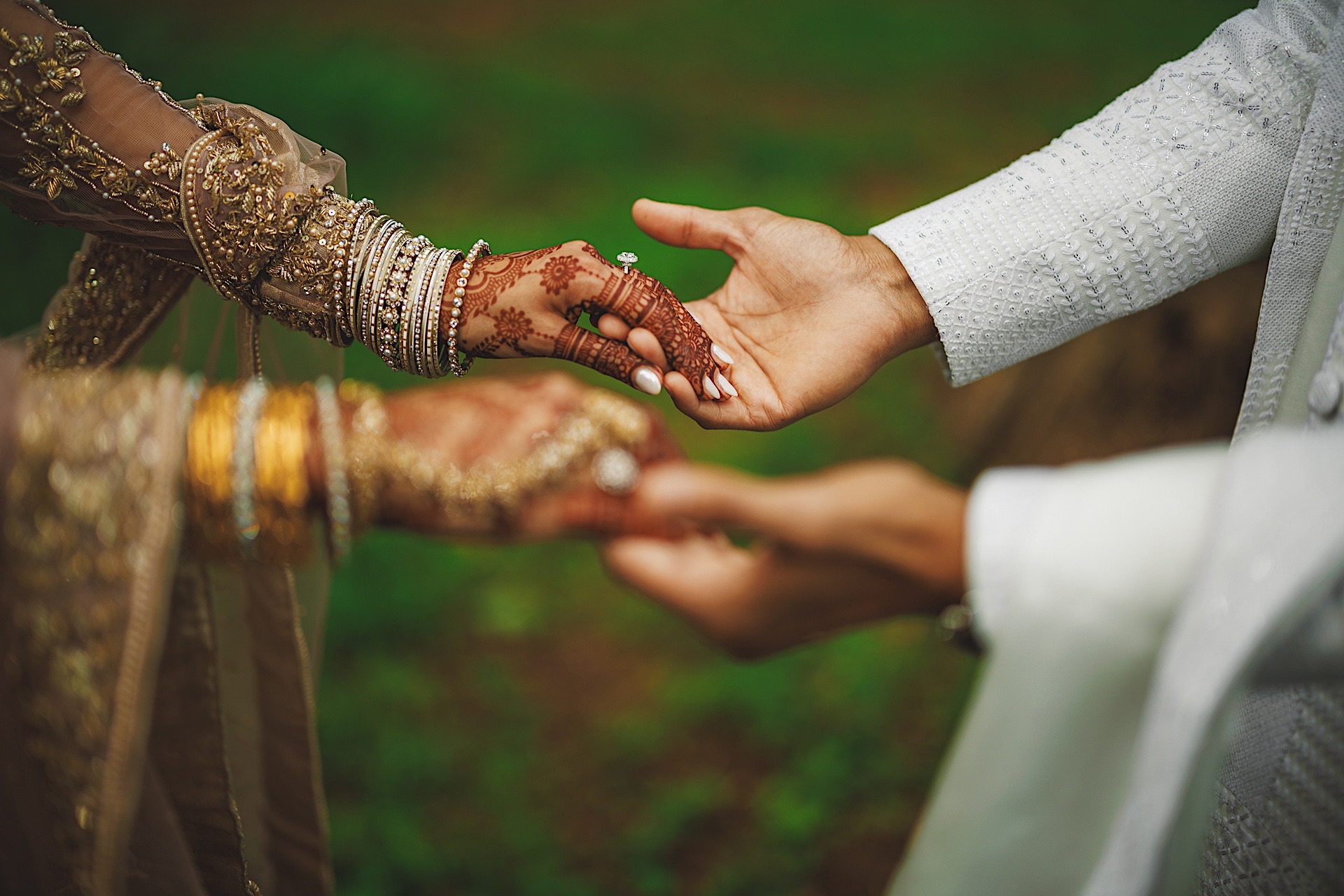 A bride and groom wearing traditional ethnic clothing hold hands, with intricate henna designs visible on the bride's hands. Set against a lush green outdoor background, their joy seems almost like an Auto Draft of a perfect moment.