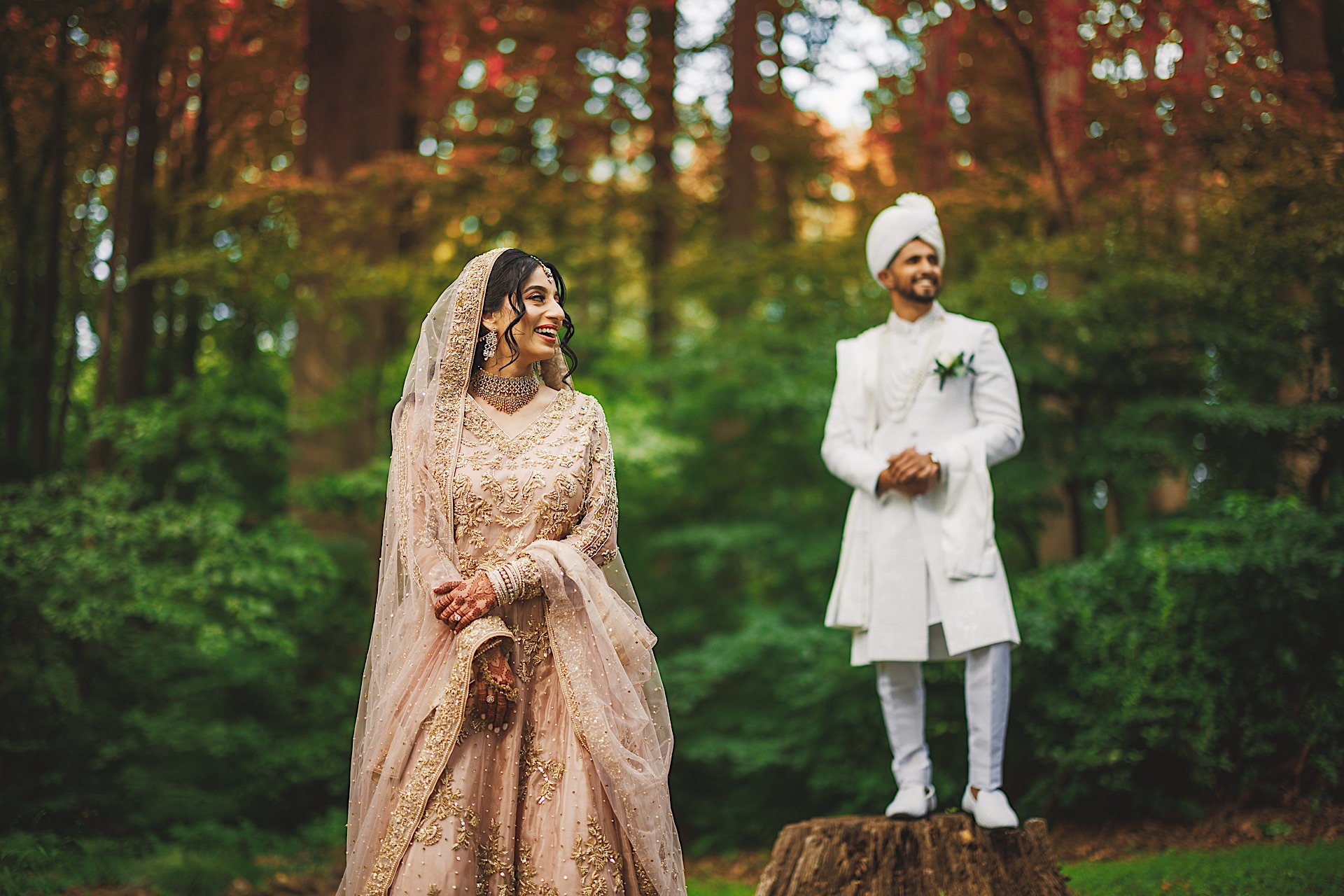 A bride in a traditional dress smiles while standing in a forest, and a groom in white attire and turban stands confidently on an old tree stump behind her, like an auto draft of nature’s artistry.