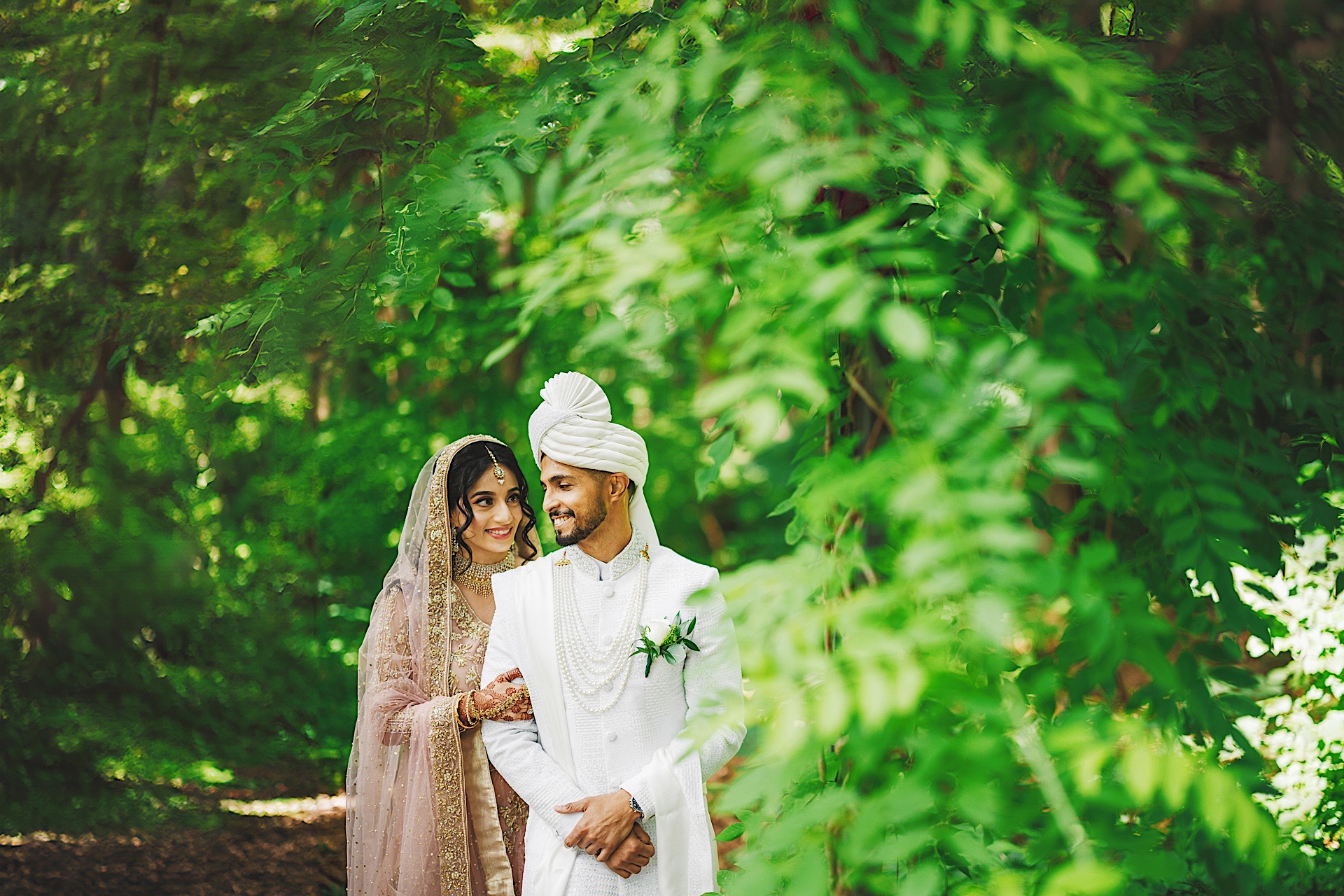 A couple dressed in traditional wedding attire stands together outdoors, surrounded by lush green foliage. The man wears a white sherwani and turban, while the woman wears a decorated headscarf and dress—each detail meticulously planned to avoid any auto draft mishap.