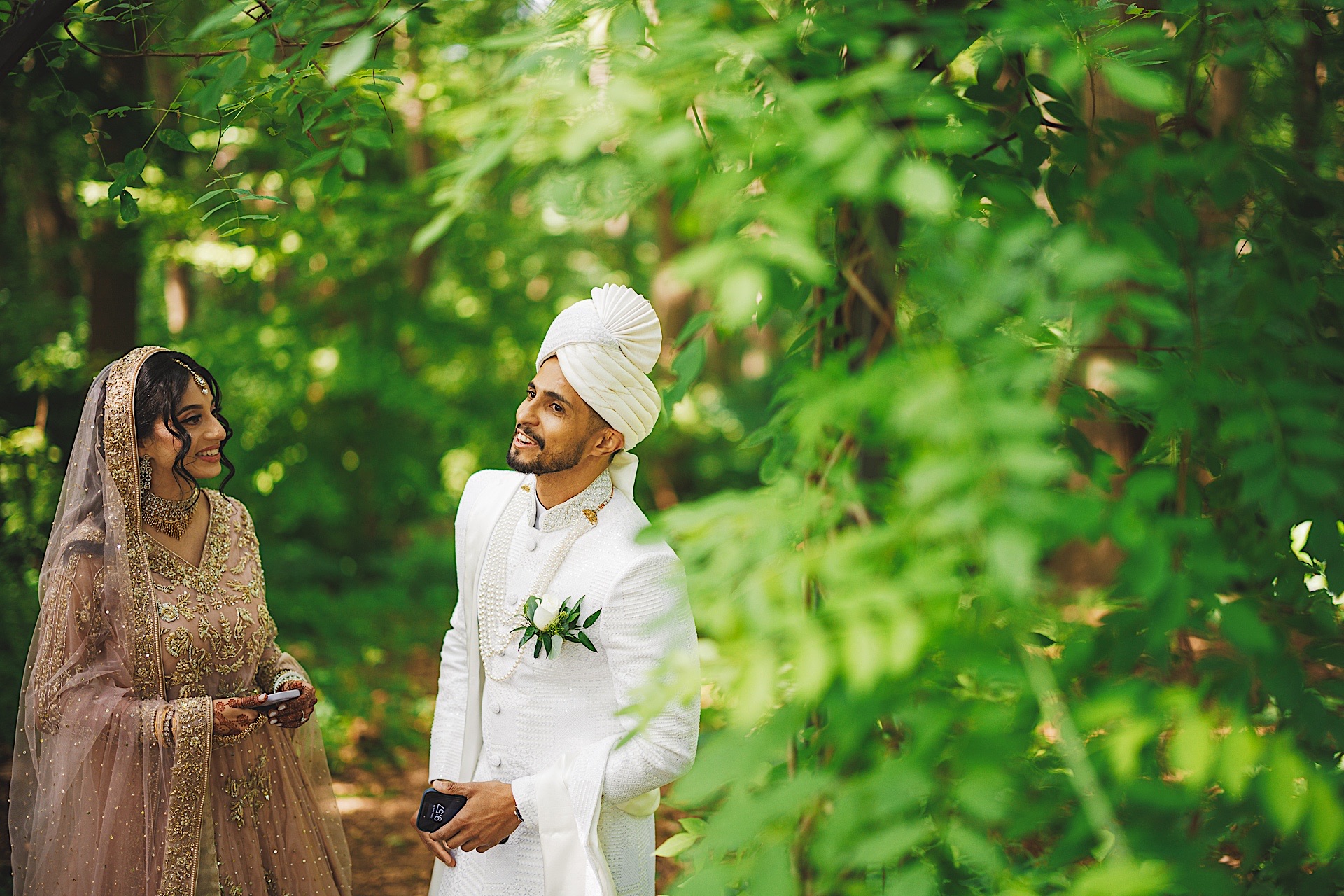 A bride and groom in traditional wedding attire stand together in a lush green forest, smiling and looking at each other. The bride wears a beaded dress and veil, while the groom dons a white sherwani and turban, as if straight out of an Auto Draft from a storybook romance.