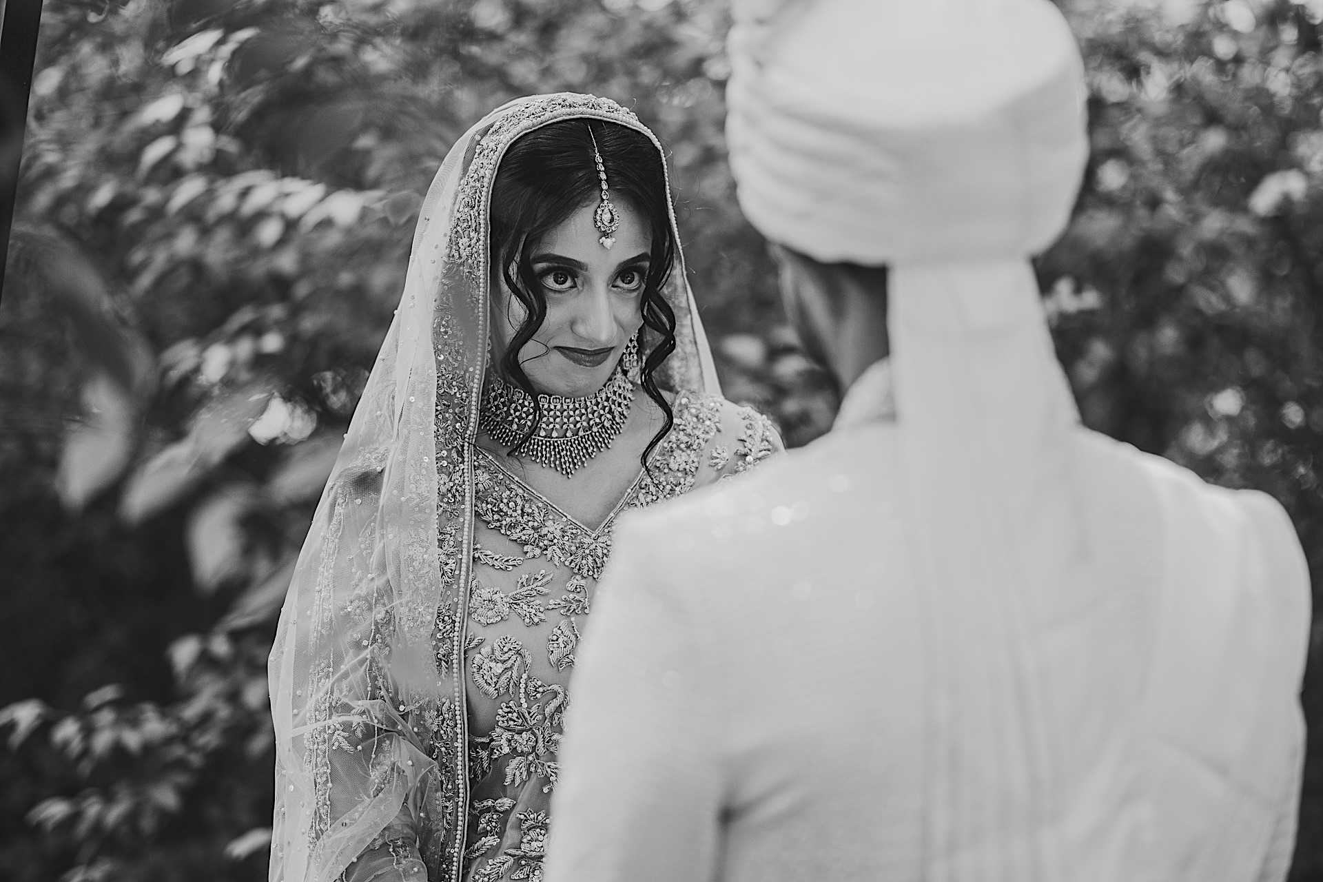 A bride, adorned in traditional attire and a veil, holds a gaze with a groom in a turban during their wedding ceremony. This captivating black and white photo, with its blurred nature background, seems like an auto draft of timeless love.