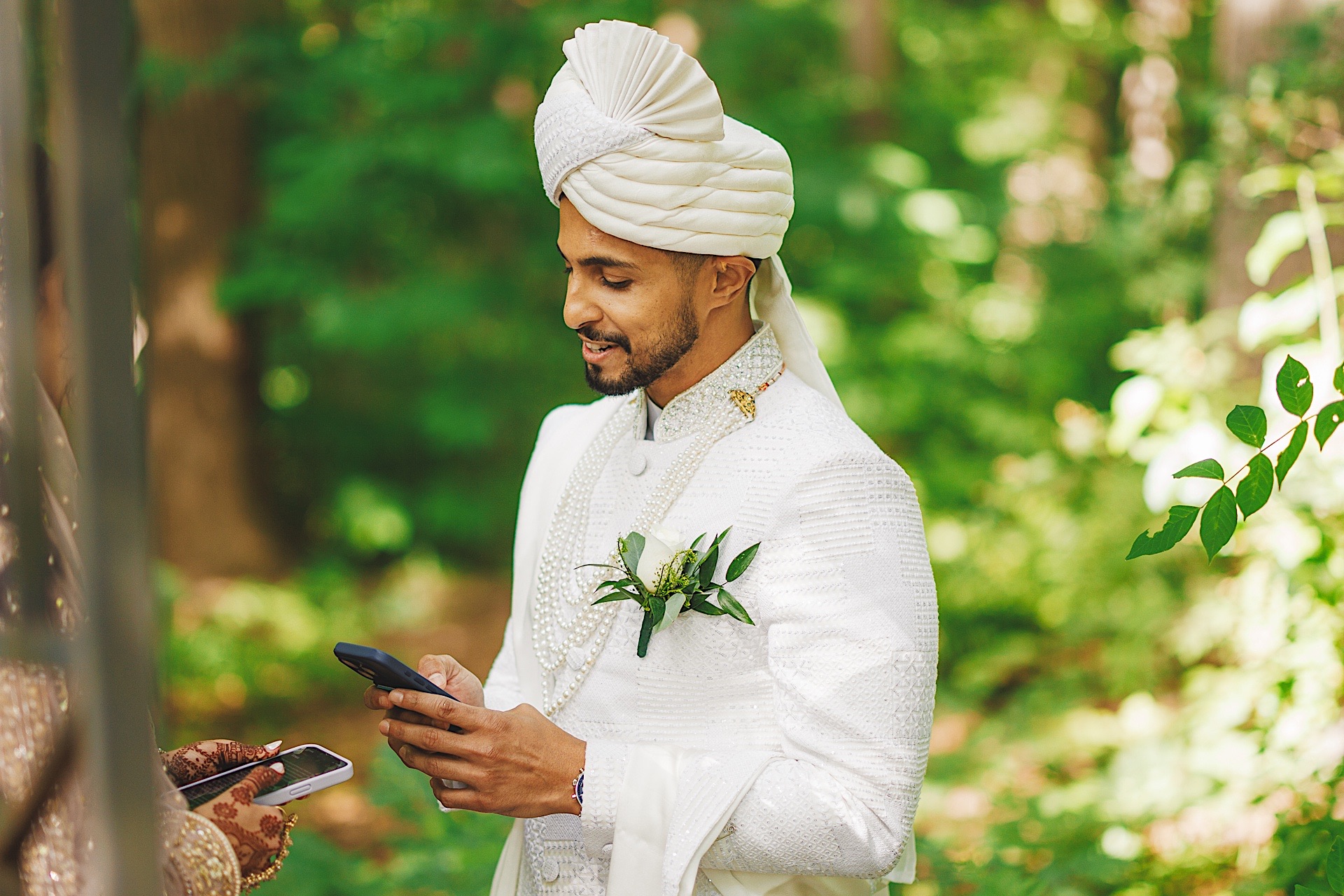 A man in a white traditional outfit and turban holds a smartphone while standing outdoors. He has a boutonniere pinned on his jacket and appears to be reading or drafting something important.