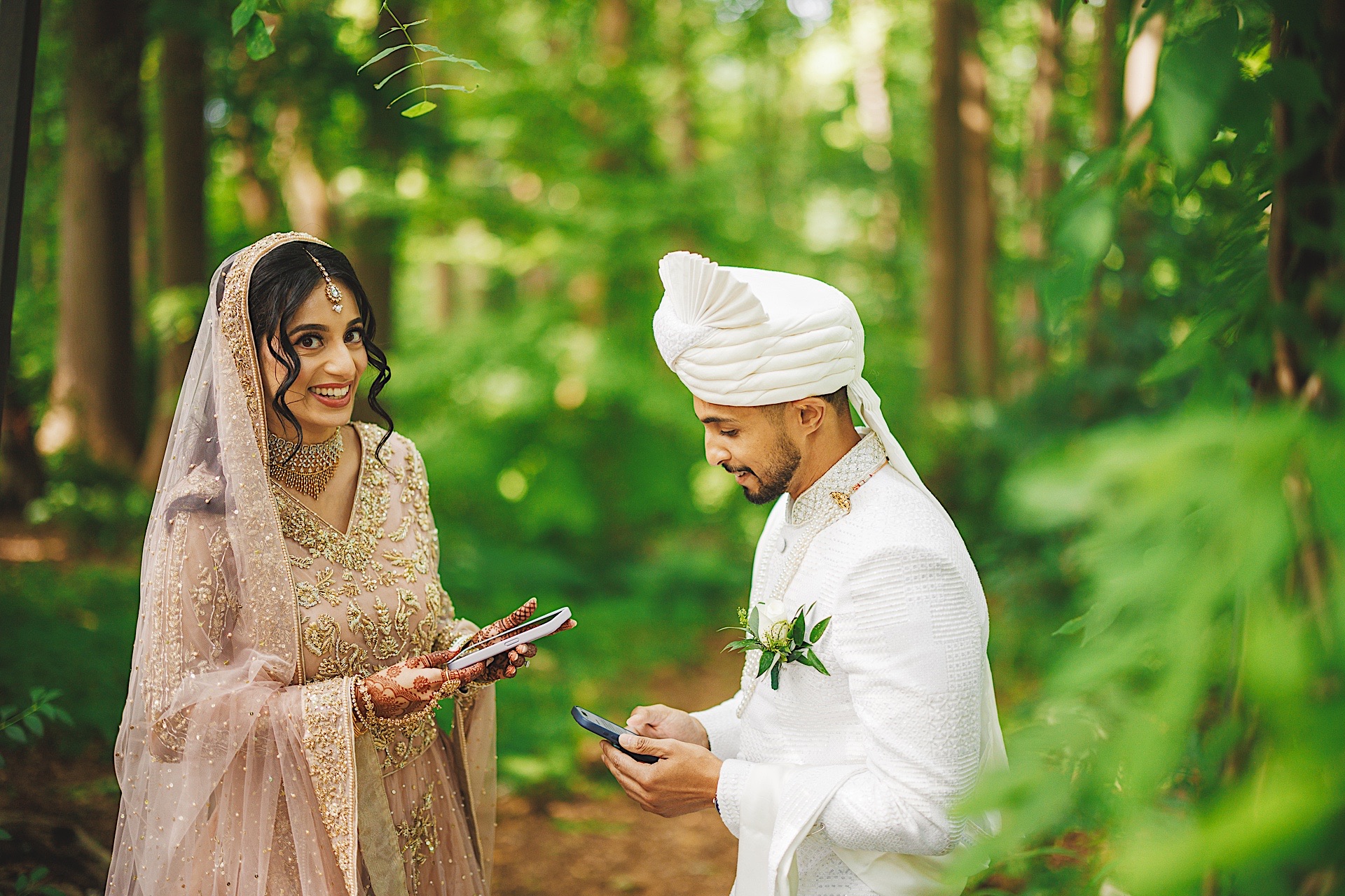 A bride and groom stand in a forest exchanging smiles and holding their phones, almost as if they are the auto drafts of a modern fairy tale. The bride wears a light pink embellished outfit, while the groom dons a white ensemble with a turban.