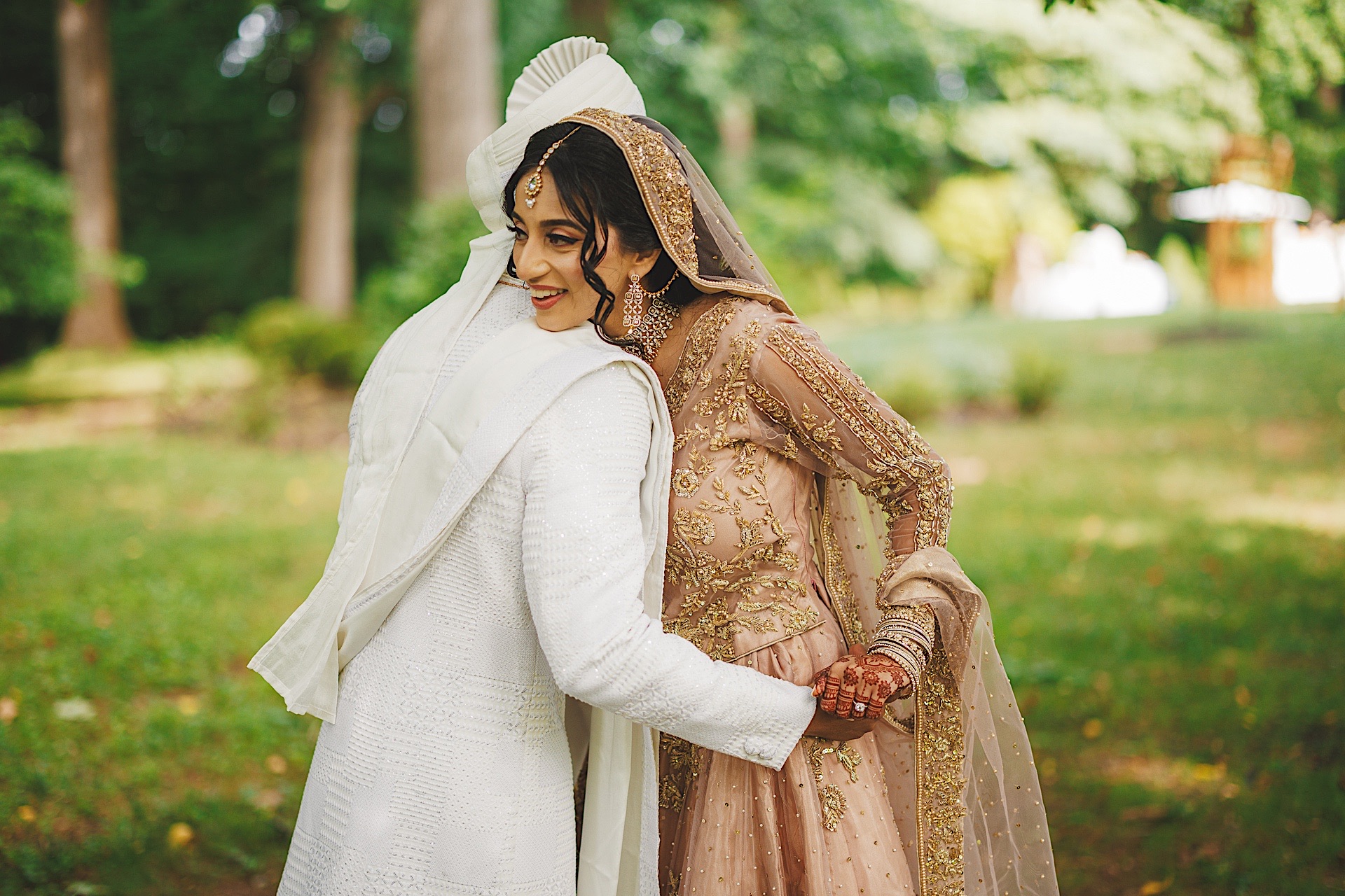 A couple dressed in traditional wedding attire embraces outdoors, as if captured in an auto draft moment. The groom wears white while the bride wears an ornate beige outfit with a matching head scarf. Greenery is visible in the background.